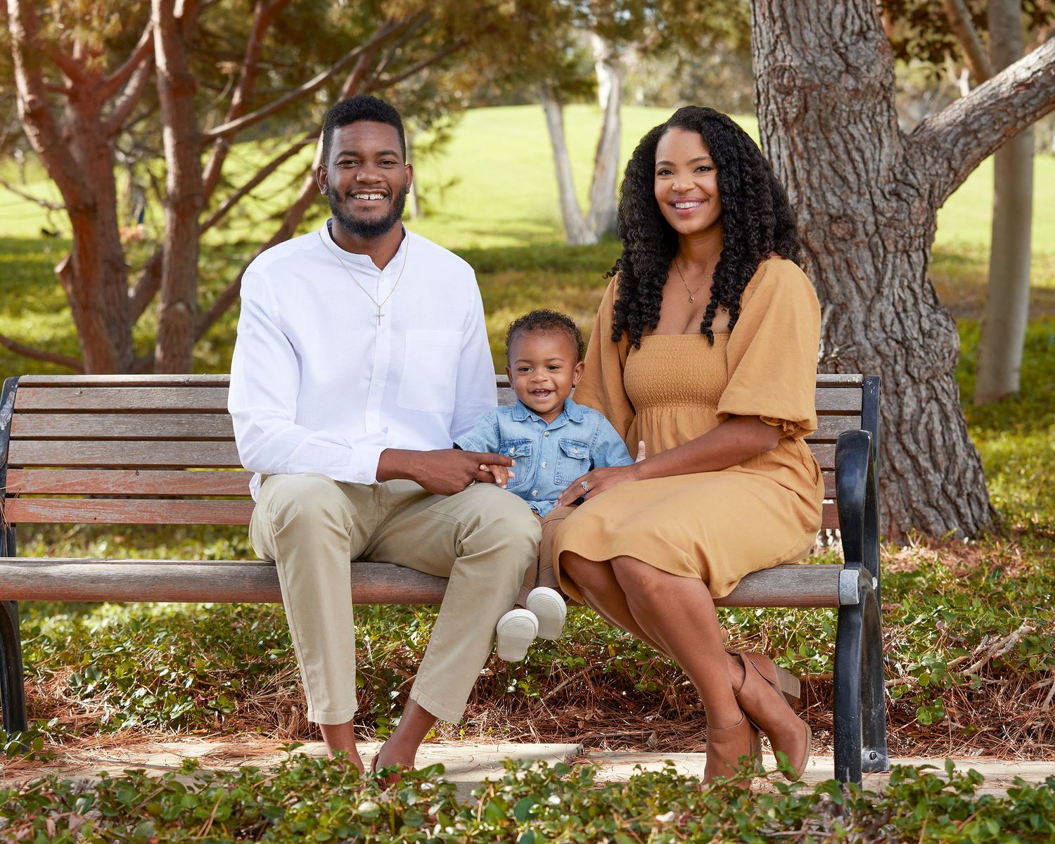 A family sits together on a wooden park bench under trees on a sunny day.
