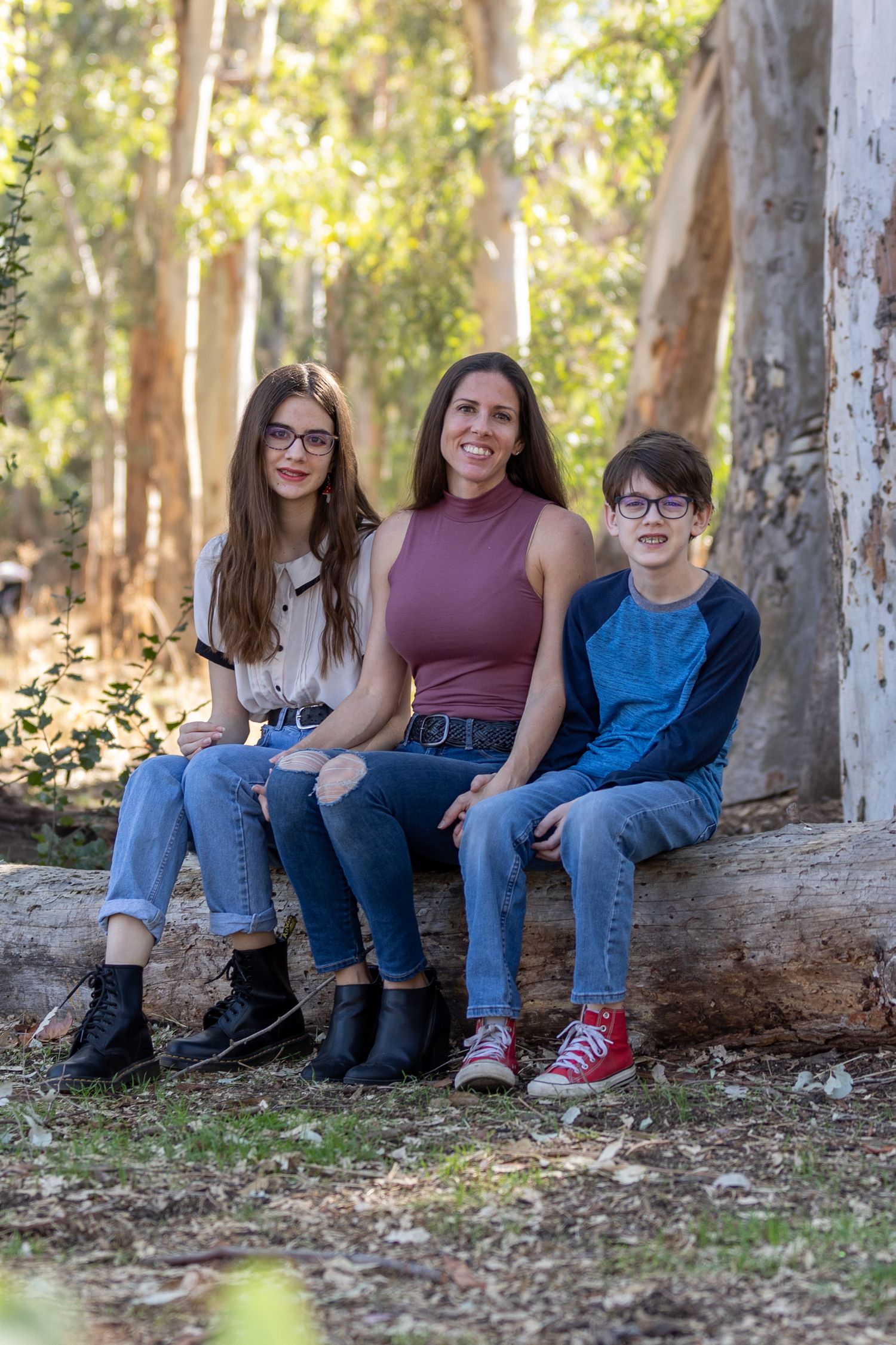 A family of three sits together on a fallen tree trunk in a sun-dappled eucalyptus grove.