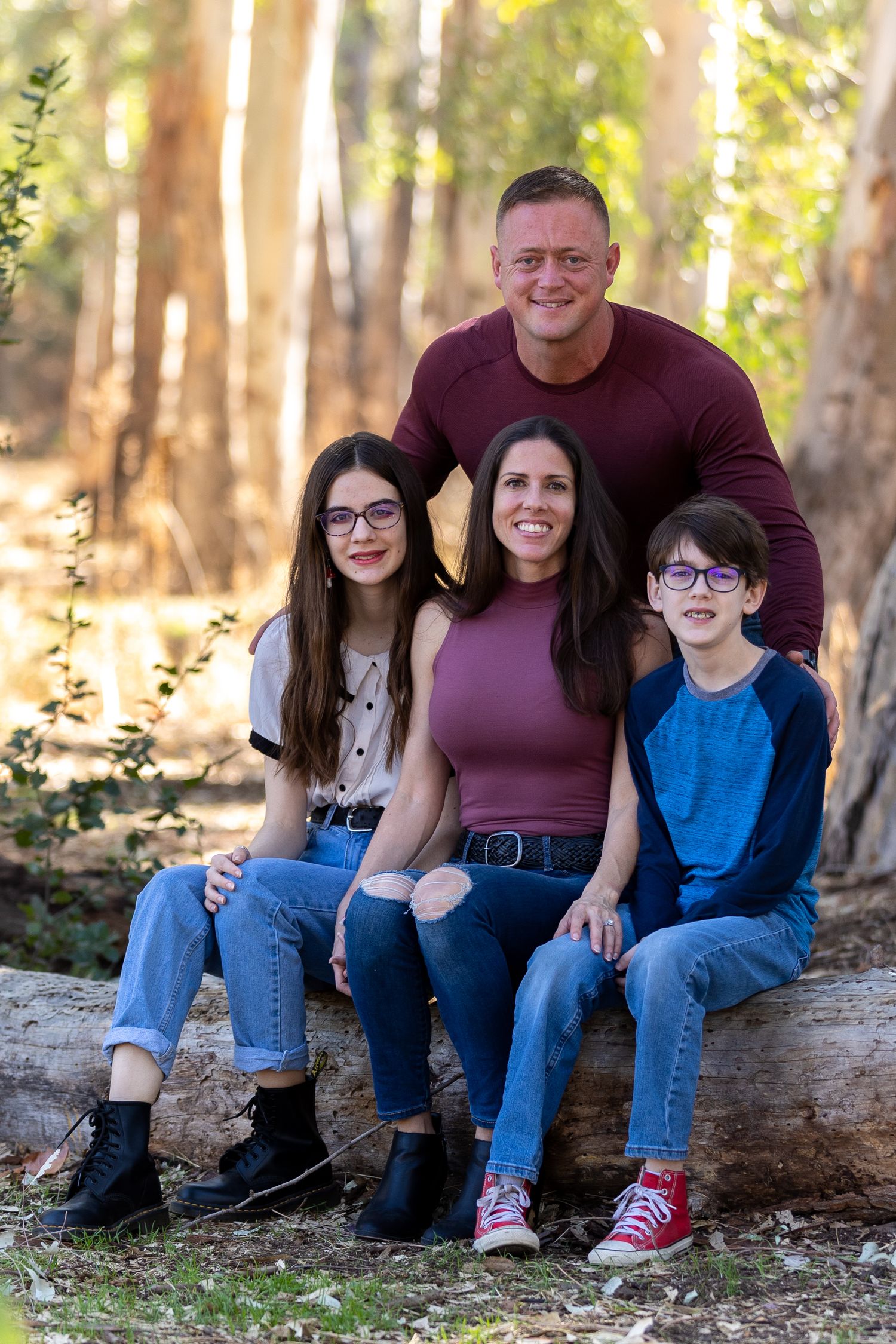 A family of four poses together while sitting on a fallen tree in a sunlit forest.