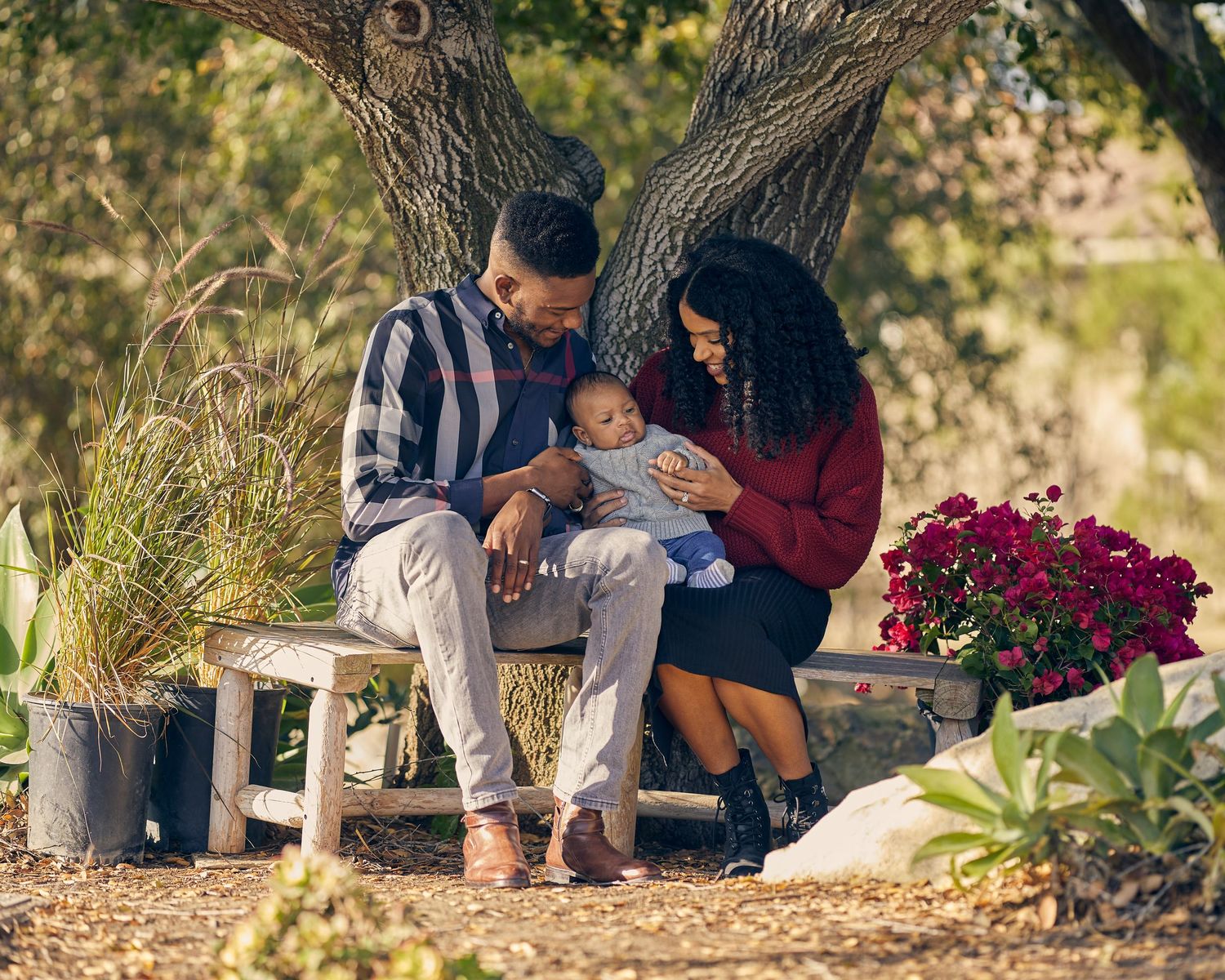 A couple shares a tender moment with their baby while sitting on a garden bench surrounded by flowers.