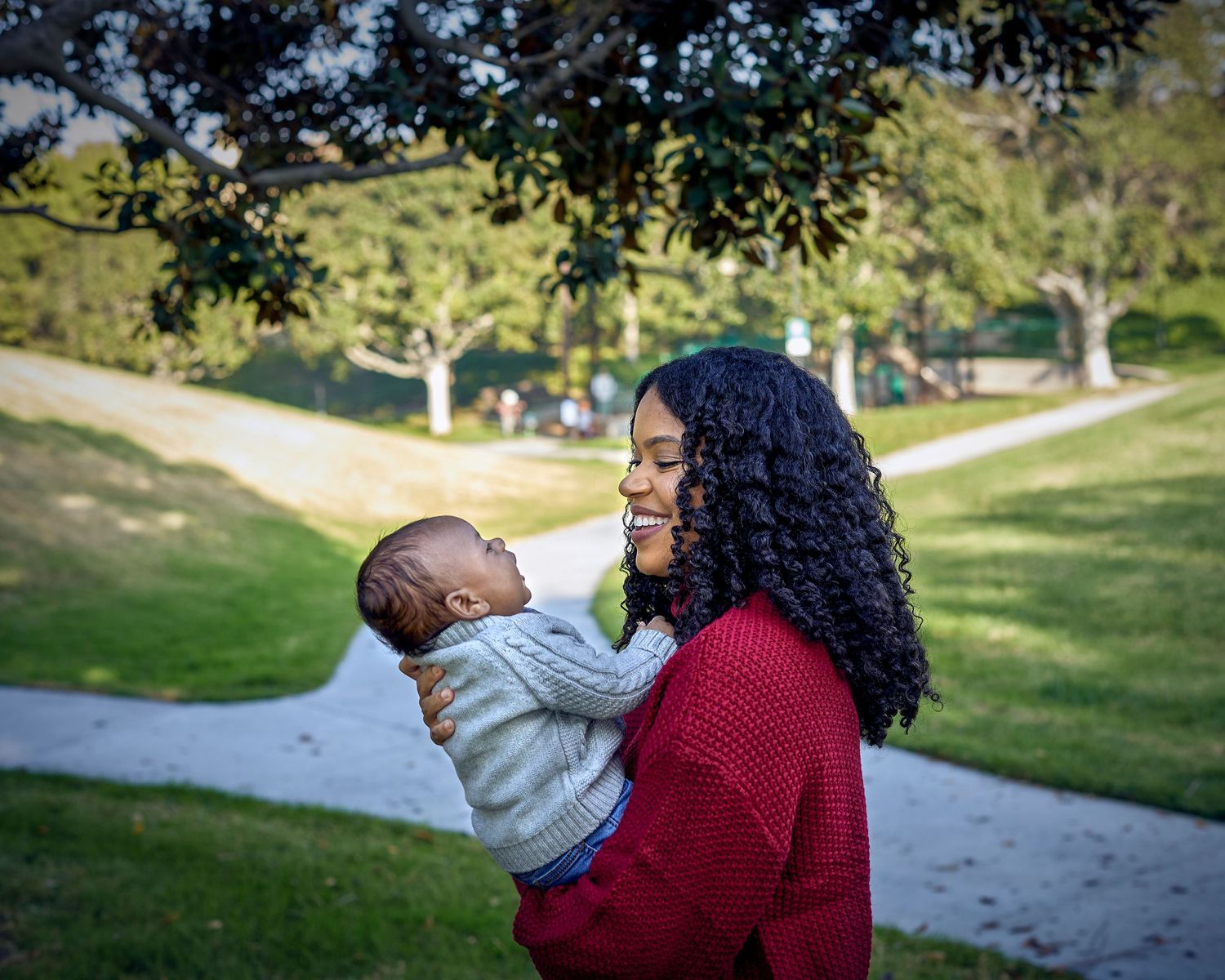 A parent holds a baby while standing in a sunny park setting.