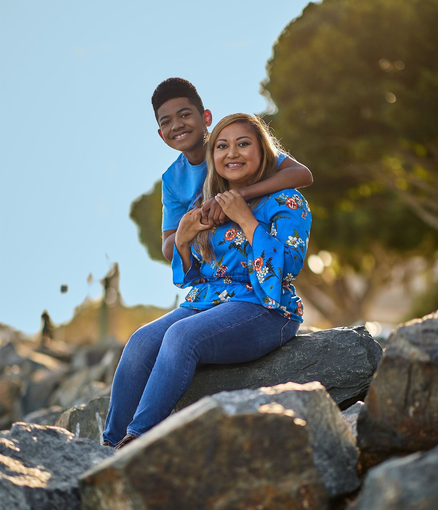 A couple sits on rocky outcrop sharing a joyful moment in warm sunlight while wearing casual attire.