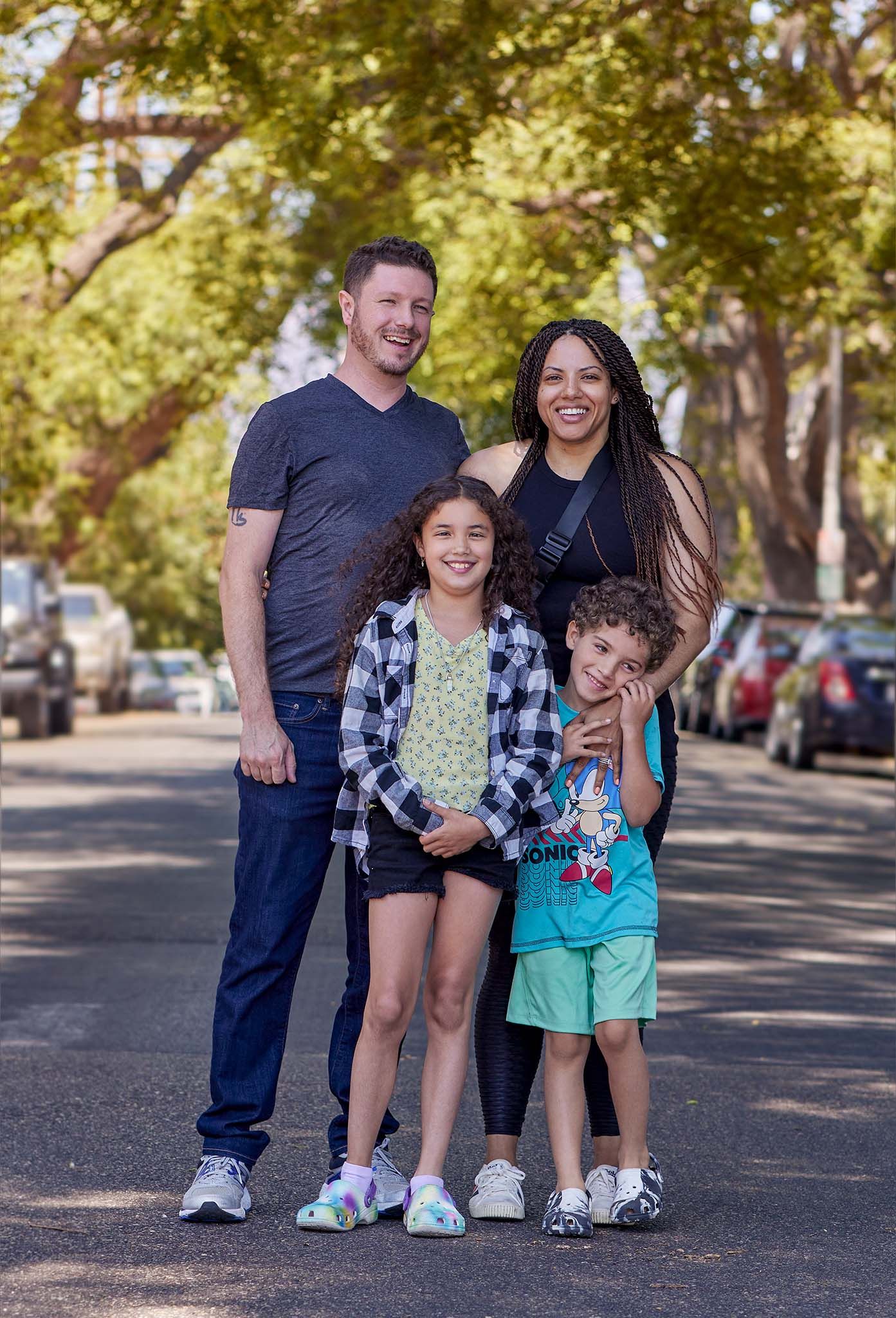 A happy family of four poses together on a tree-lined street.