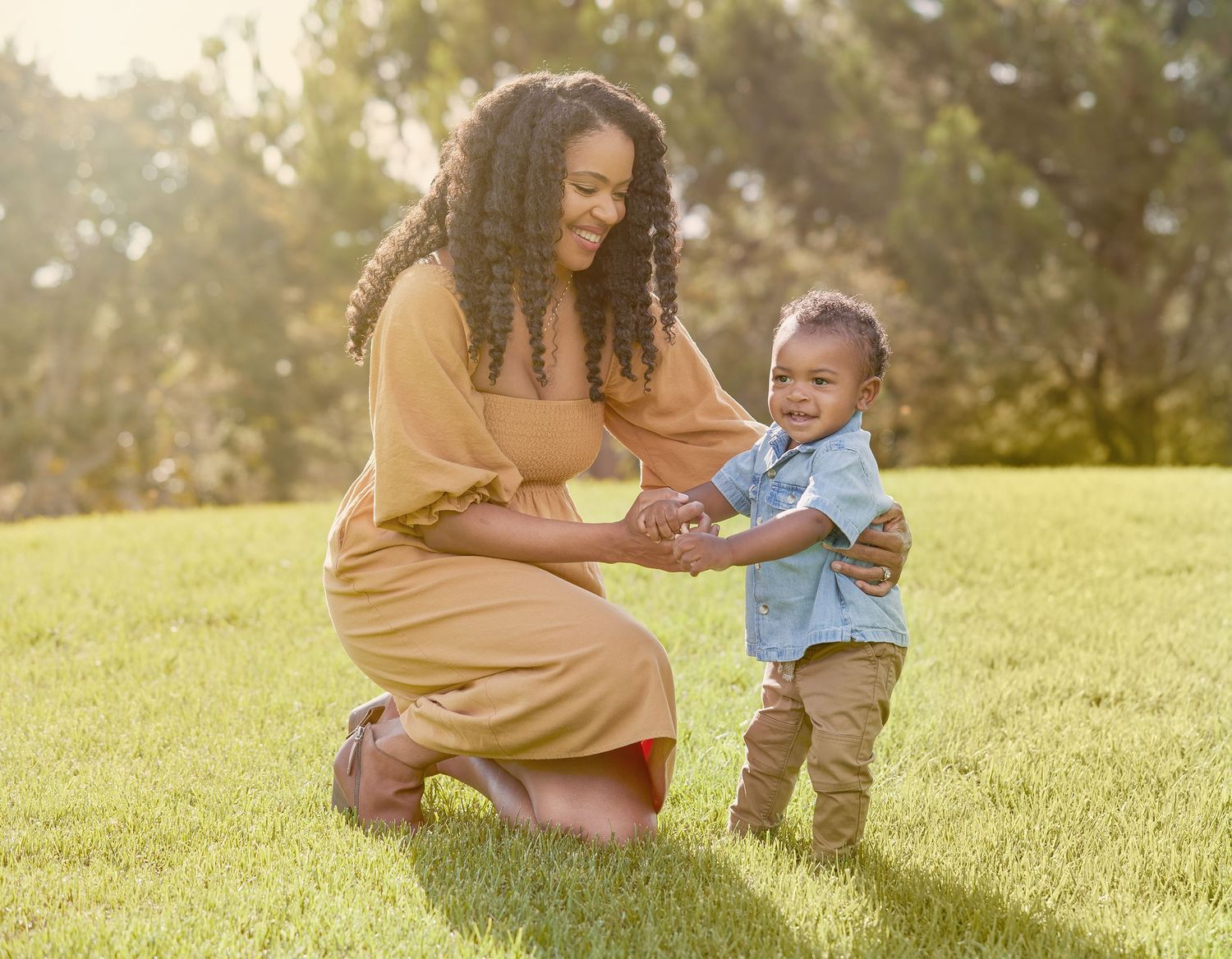A toddler in denim plays with a caregiver on lush green grass in a park setting.
