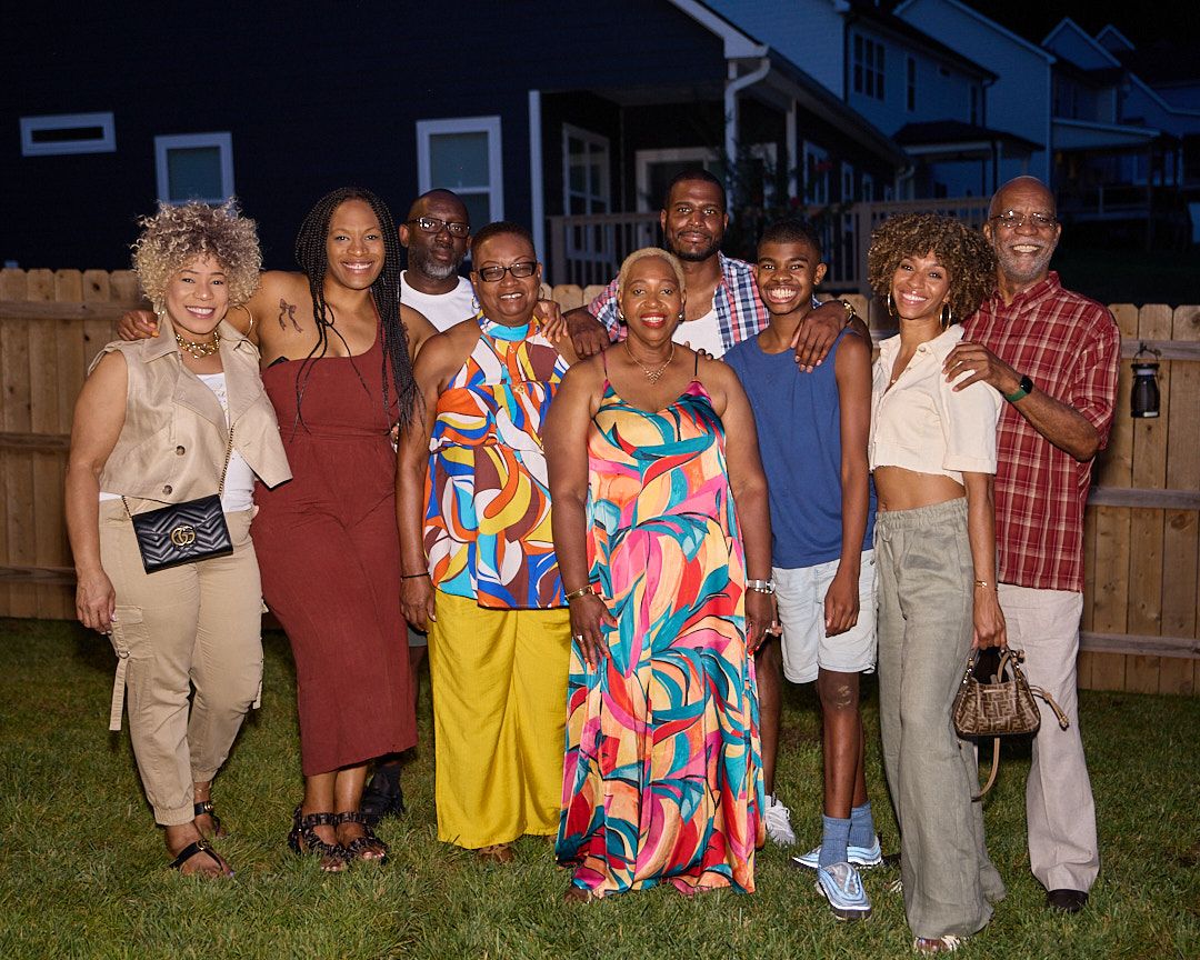 Family gathering photo with multiple people wearing colorful outfits in a backyard setting.
