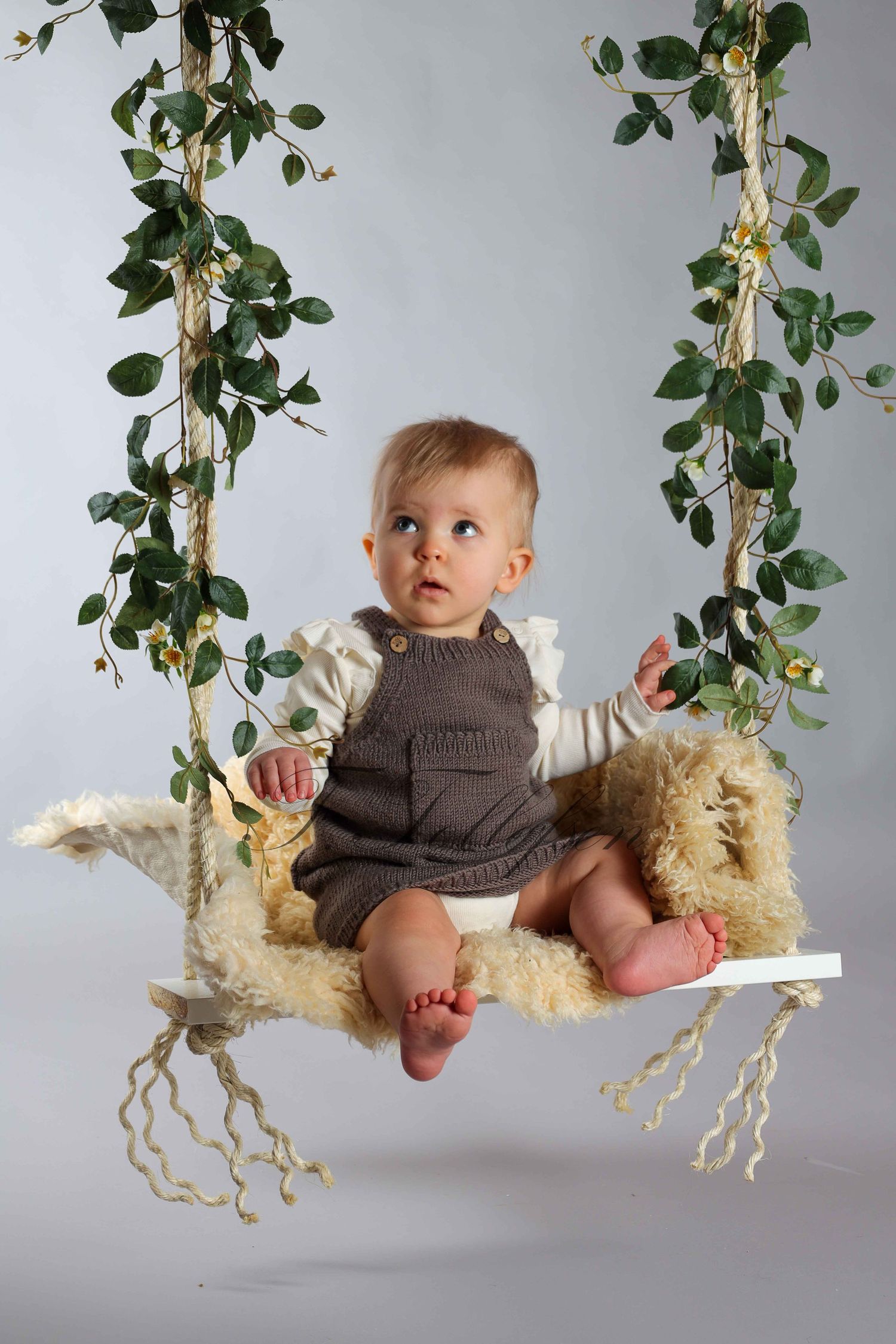 Adorable baby sitting on a fluffy swing decorated with green ivy vines in a studio photo shoot.