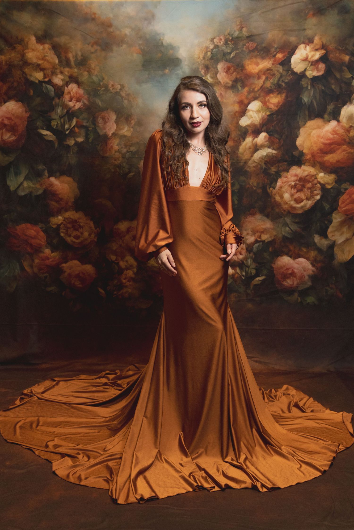 Woman in flowing copper gown poses against floral backdrop in vintage-inspired fine-art portrait. Shot by Mika Larson Photography.