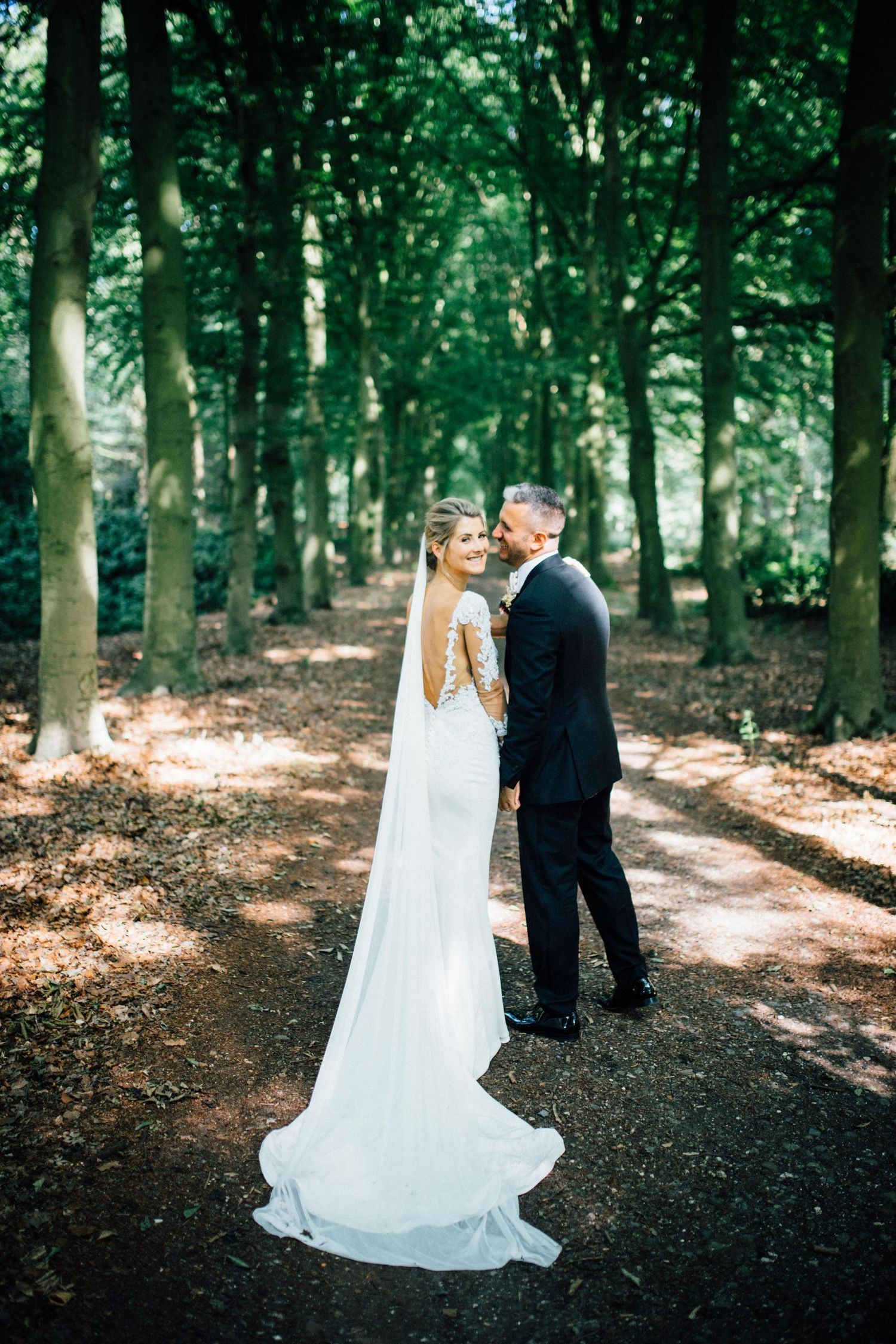 A couple in wedding attire poses on a tree-lined path in a serene forest setting. At Kasteel Maurick Vught.