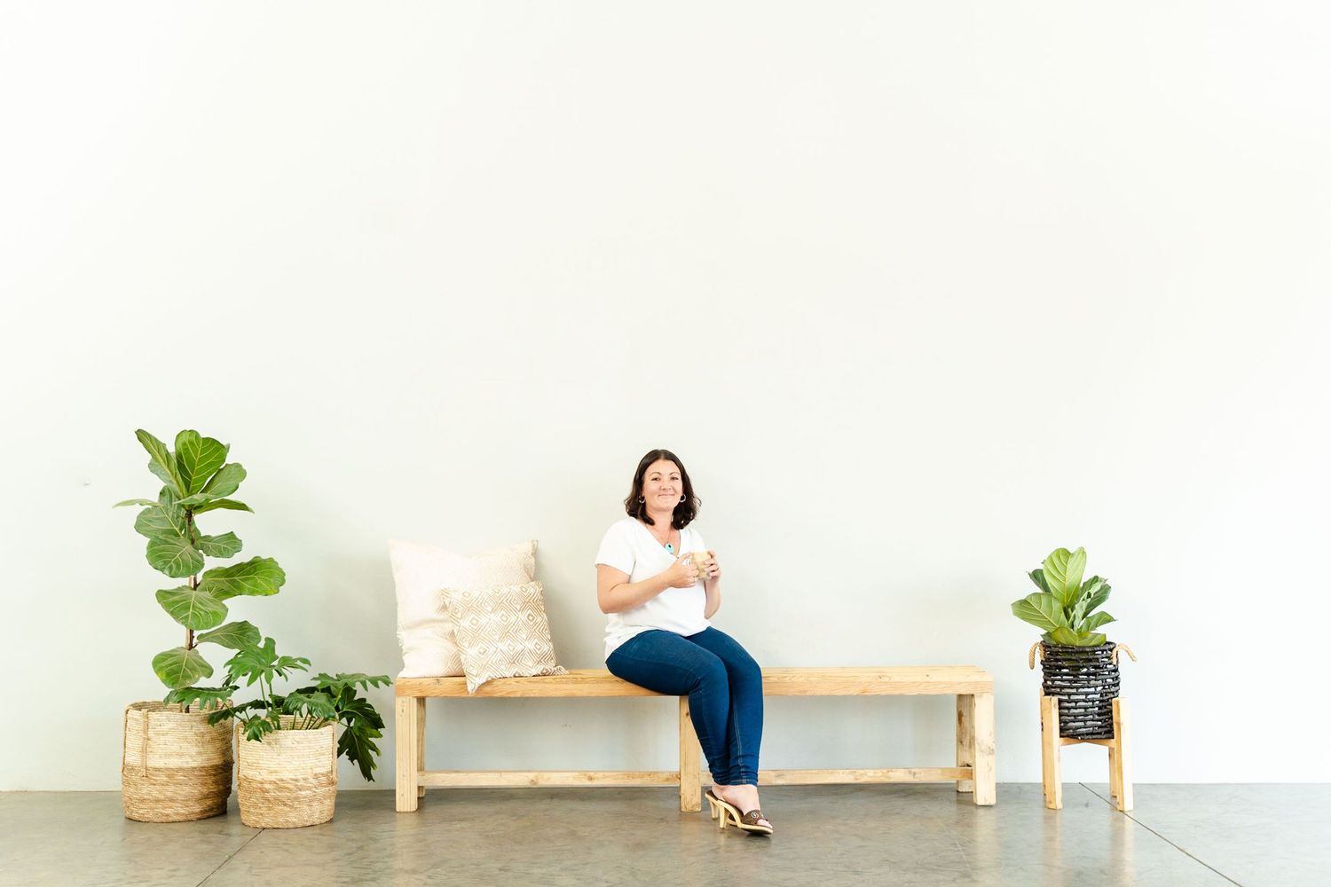Person sitting on wooden bench with throw pillows and potted plants in minimalist white room.