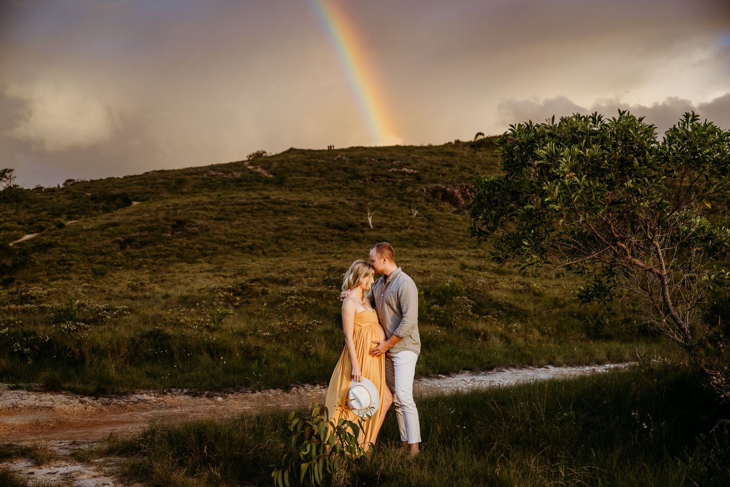 A couple embraces on a hillside path with a rainbow arching in the stormy sky behind them.