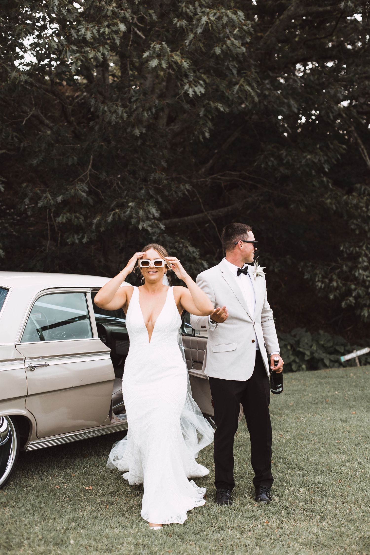 Wedding couple poses playfully in a grassy field next to a vintage car.