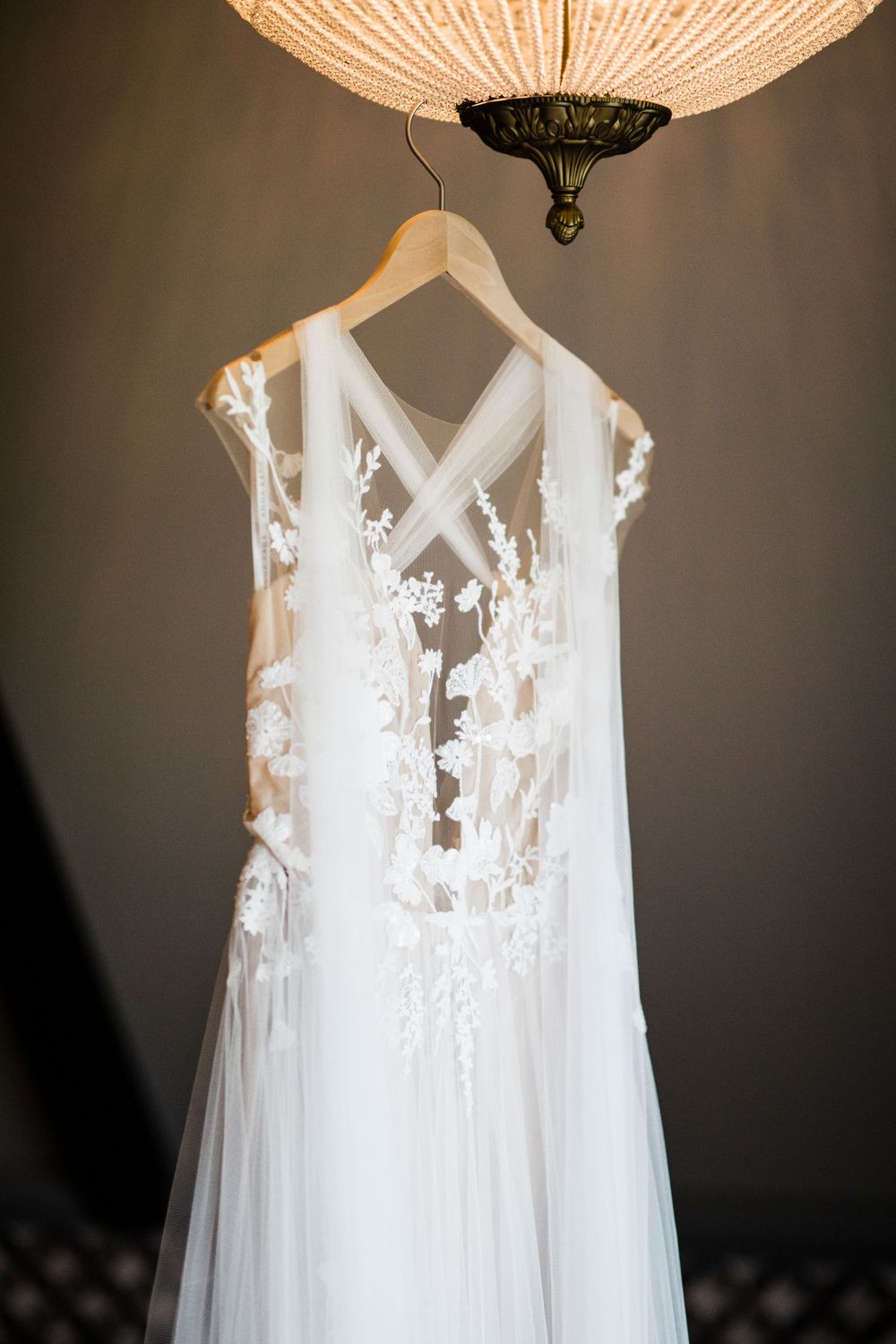 Elegant white lace wedding dress with sheer details hanging from wooden hanger under ornate chandelier.