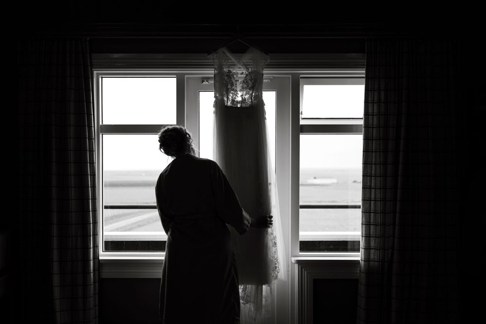 Silhouette of a figure standing by windows with a hanging wedding dress in the foreground.