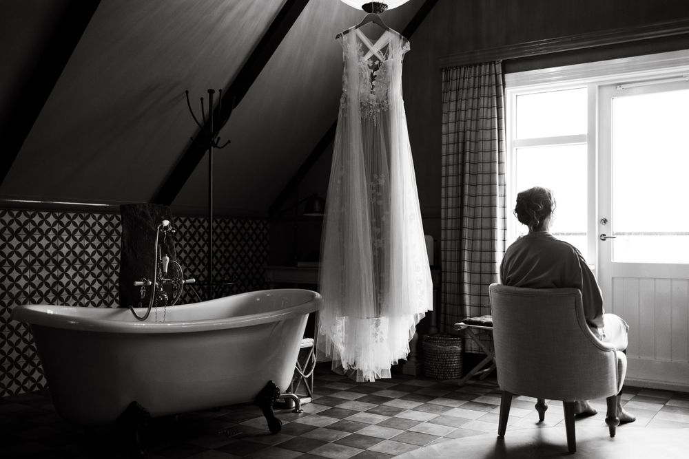 Black and white image of a wedding dress hanging in an attic room with a clawfoot bathtub.