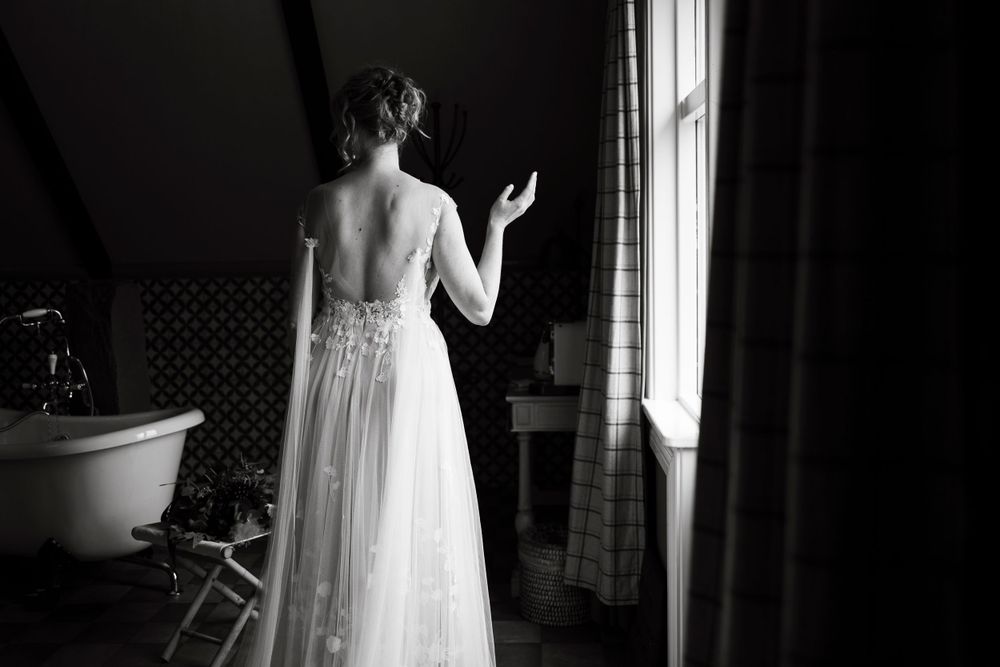 Silhouette of a bride in a backless wedding gown standing by a window in dramatic lighting.
