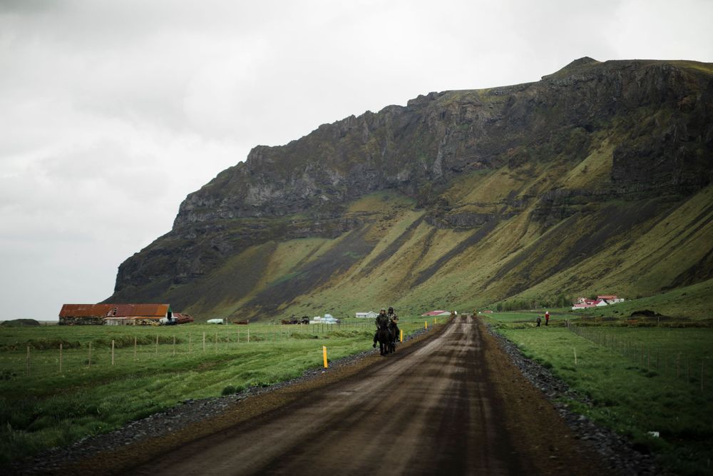 Dirt road leading through dramatic Icelandic landscape with mountains and farmhouses.