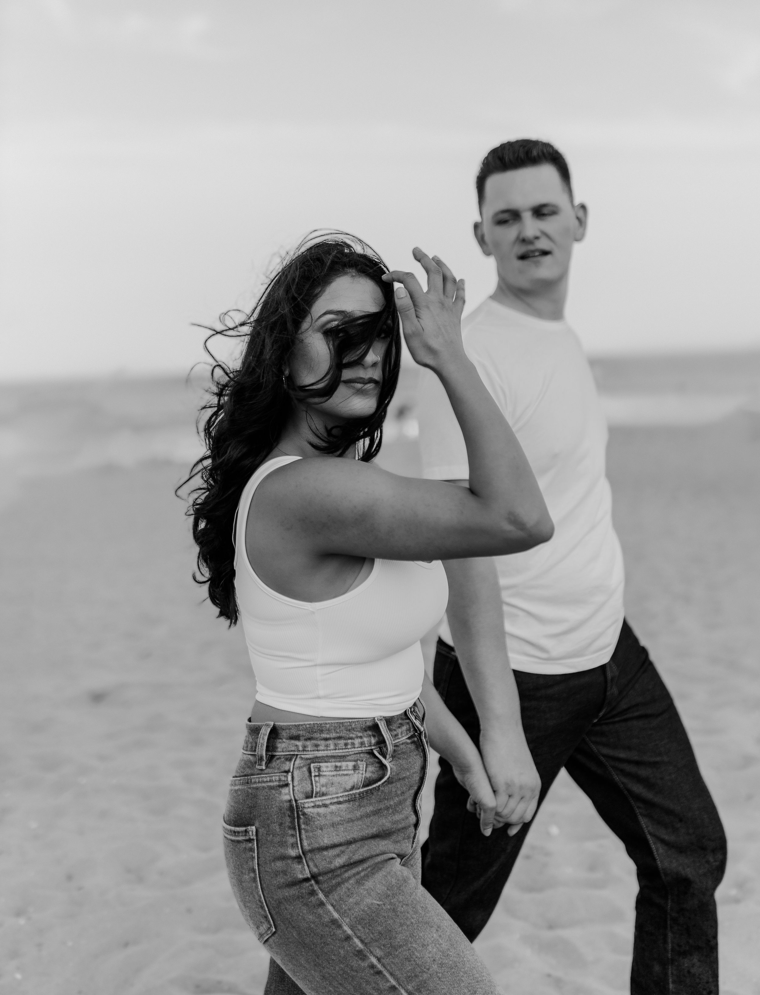 Black and white photo of a playful couple posing on a beach in casual summer outfits.