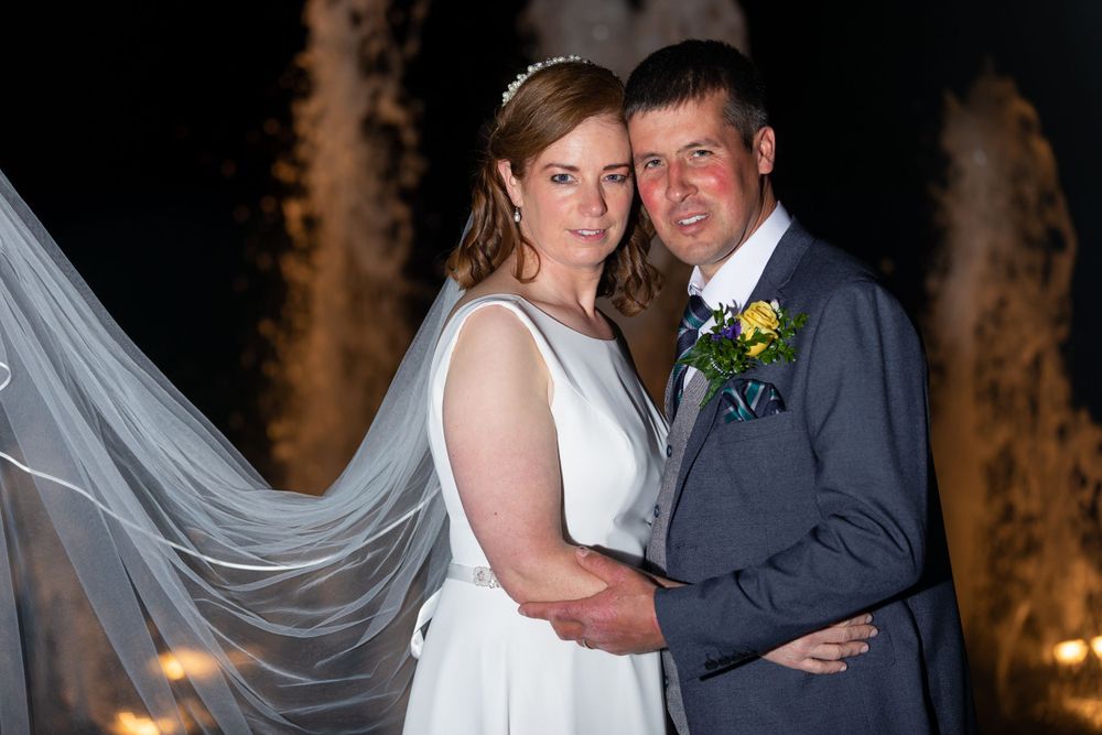 A wedding couple poses together at night with flowing bridal veil and glowing fountain backdrop.