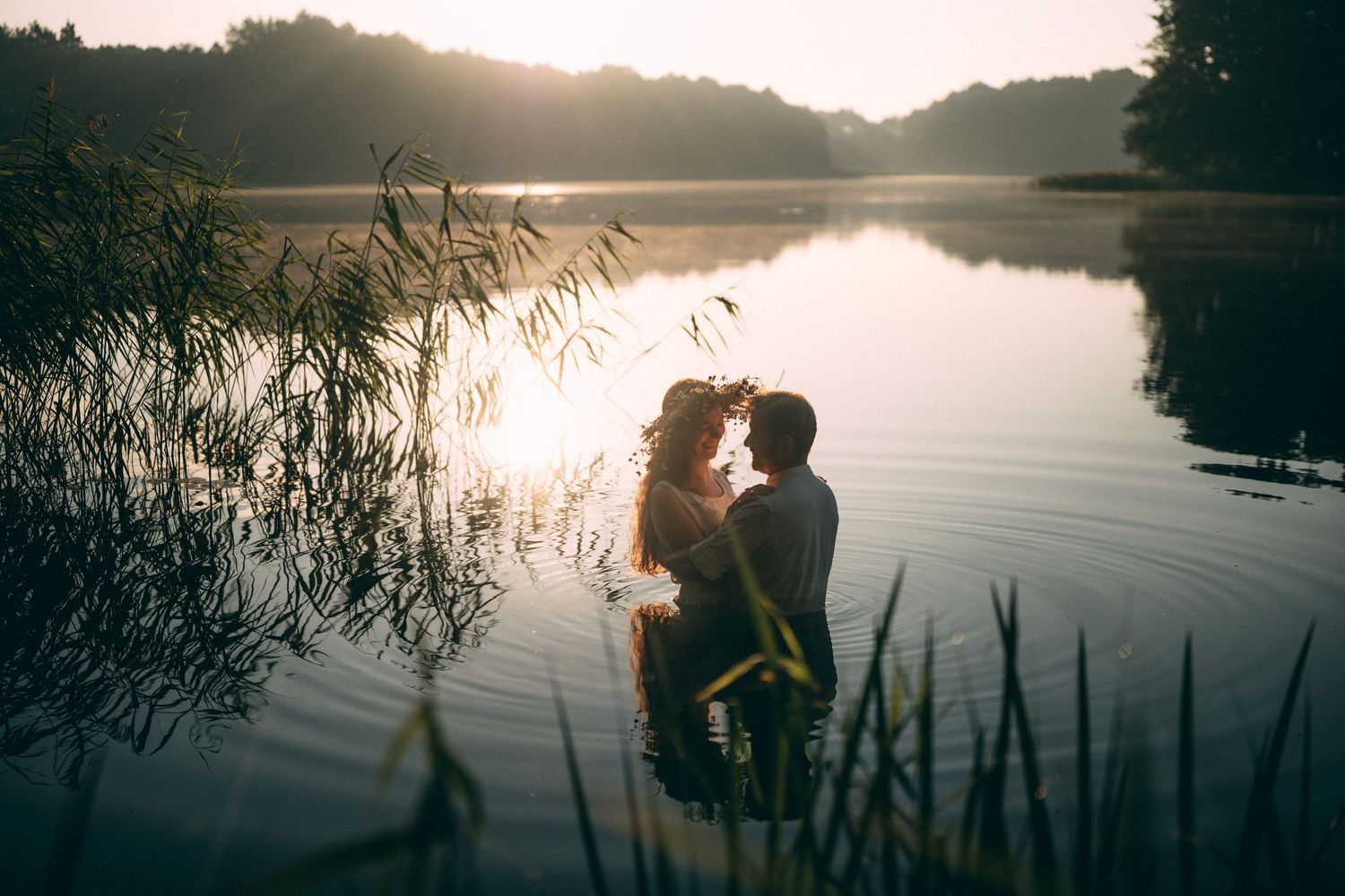 Silhouetted figures by lake at sunset with reeds in foreground.