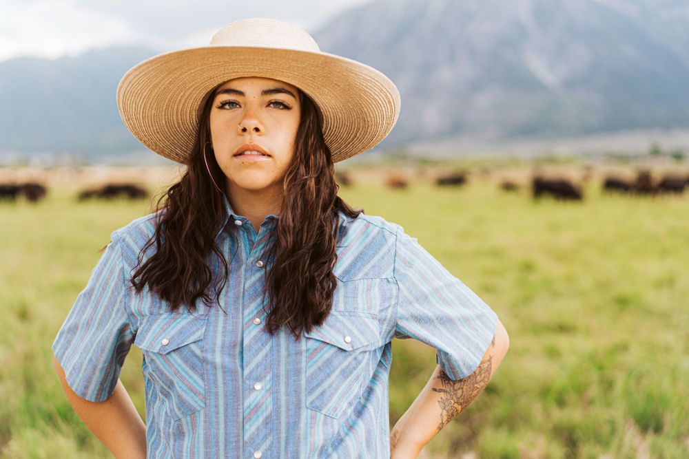 Person in western attire and sun hat poses in front of mountain landscape.