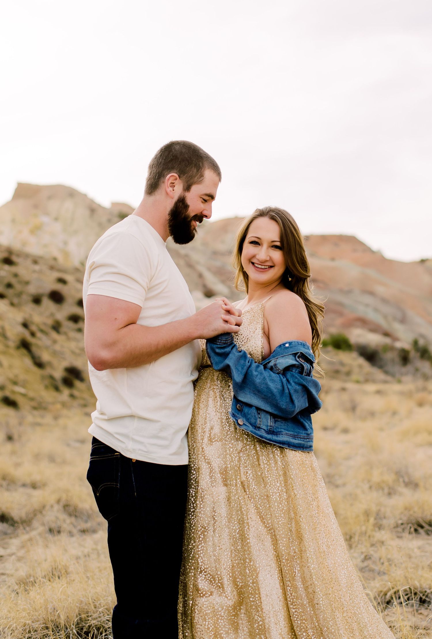 A couple shares a romantic moment against a desert mountain backdrop wearing a gold dress and casual attire.