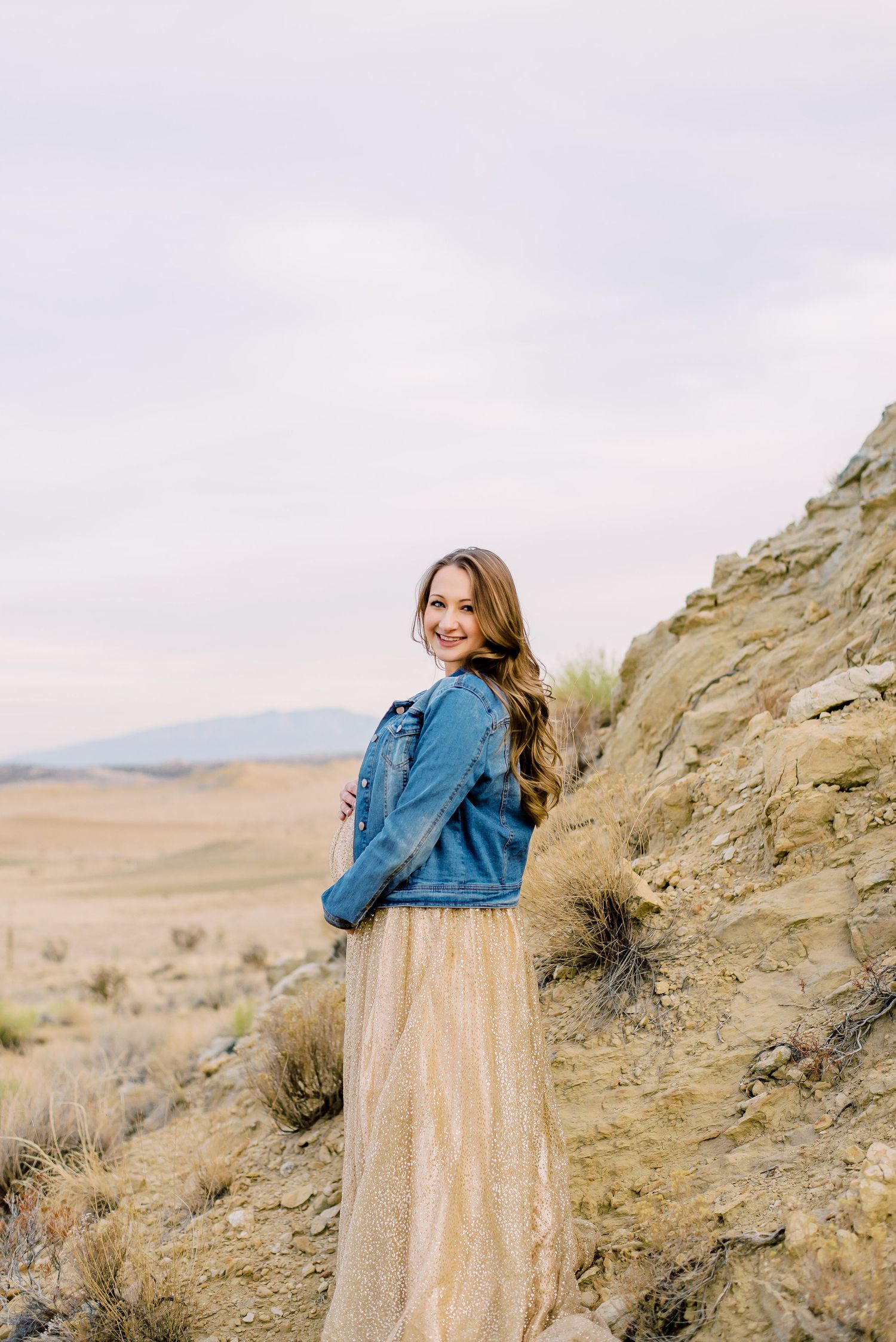 Young woman in denim jacket and gold maxi skirt smiling outdoors in a desert landscape.
