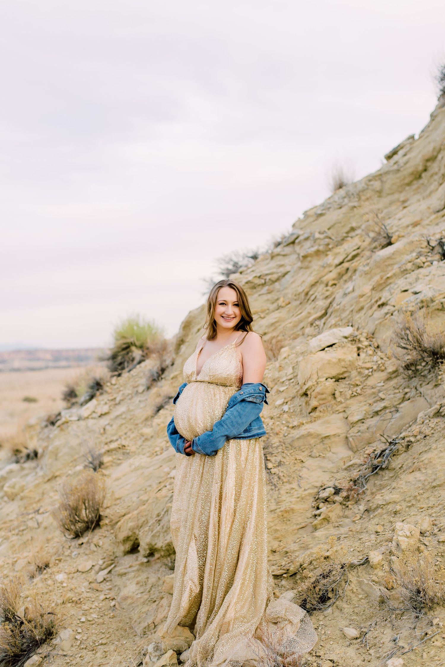 Pregnant woman in gold dress and denim jacket poses on rocky hillside during outdoor maternity photo session.