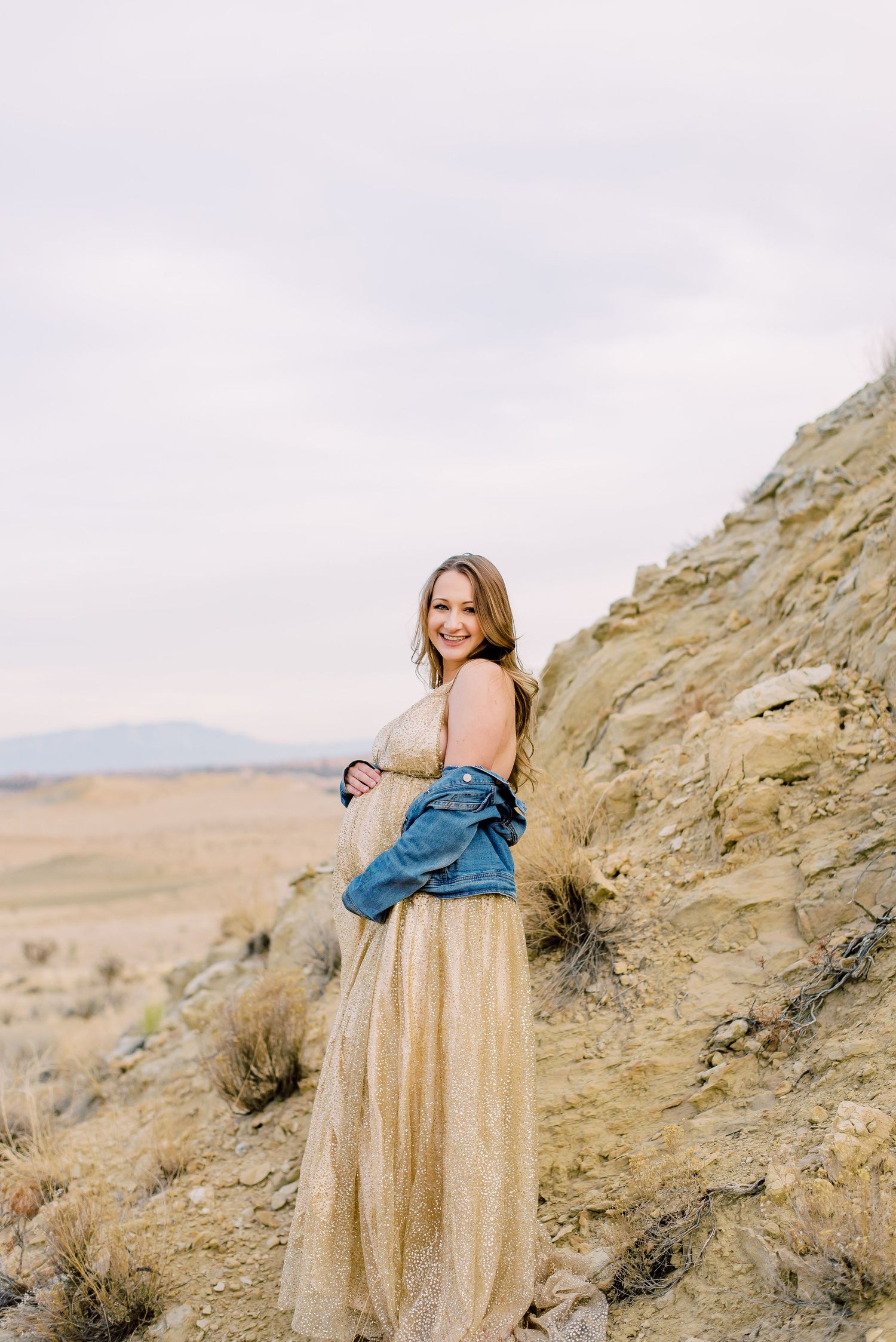 Woman in gold maxi dress and denim jacket smiling on a desert hillside with vast open landscape behind her.