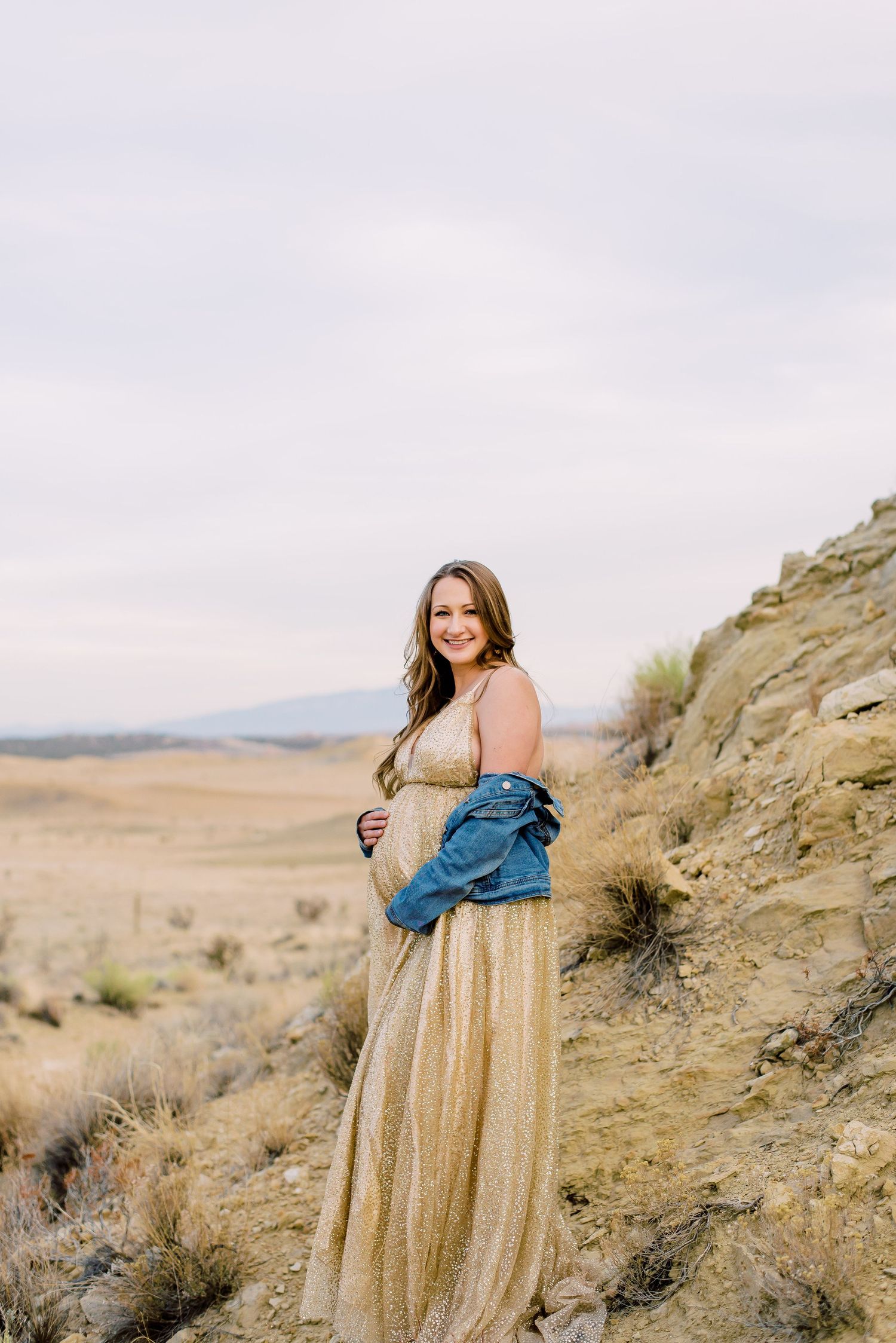 Pregnant woman in gold maxi dress and denim jacket smiling outdoors in arid desert landscape.