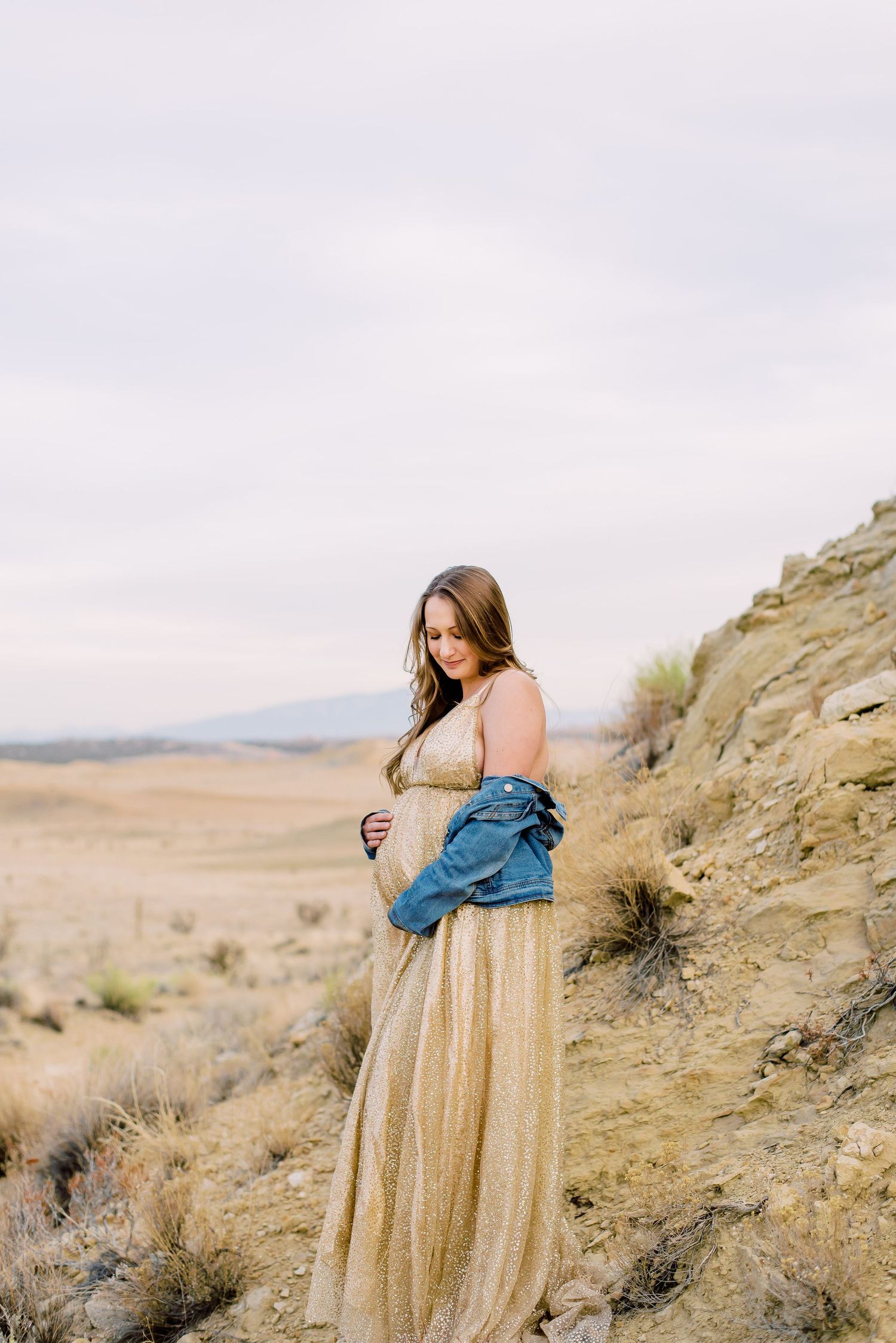 Pregnant woman in gold gown and denim jacket poses on desert hillside under overcast sky.