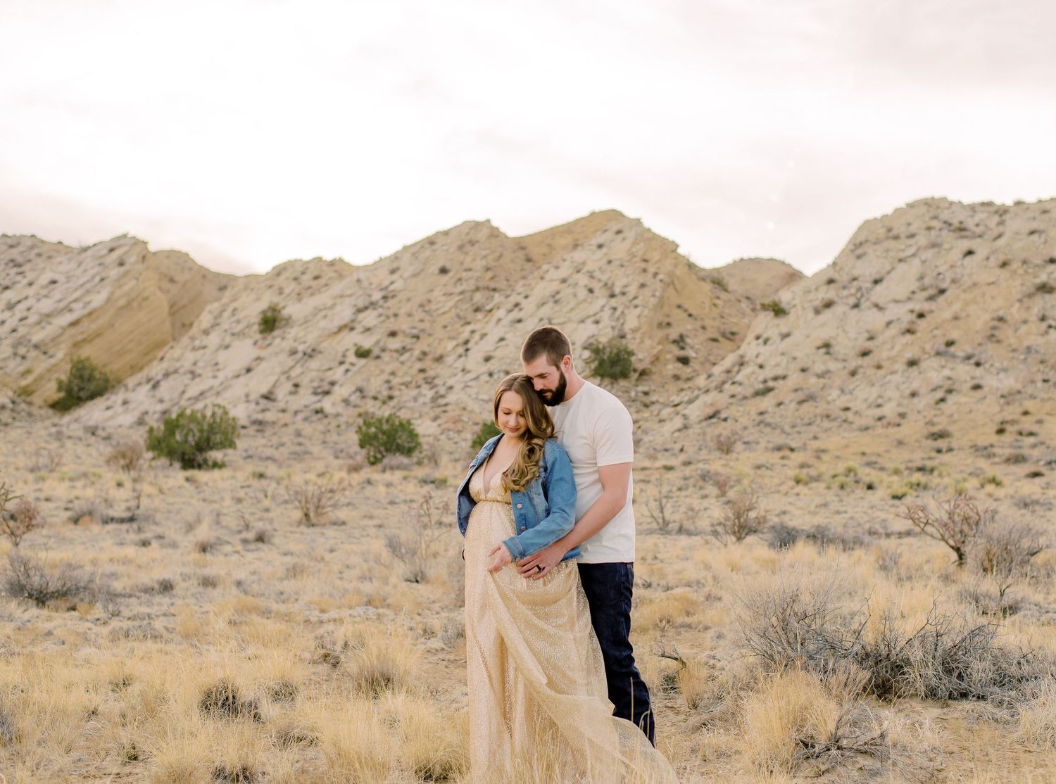 Couple embracing in desert landscape with rocky hills and dry brush in soft natural light.