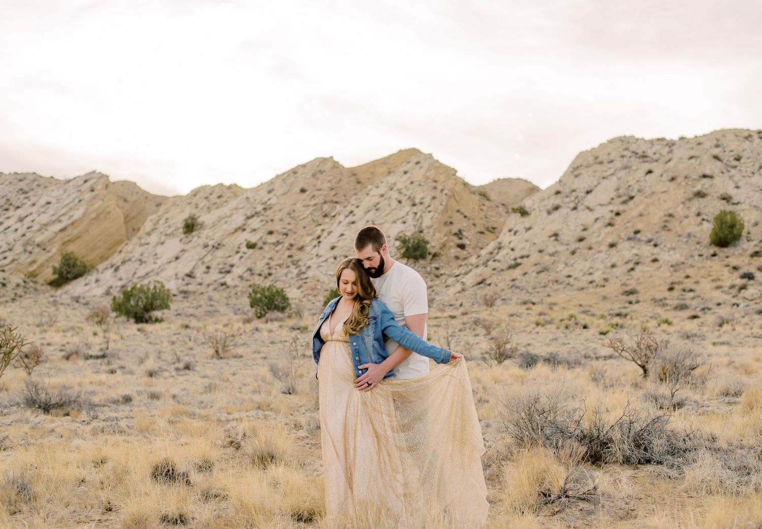 Couple embracing in desert landscape with rocky hills, woman in flowing beige gown and man in denim jacket.