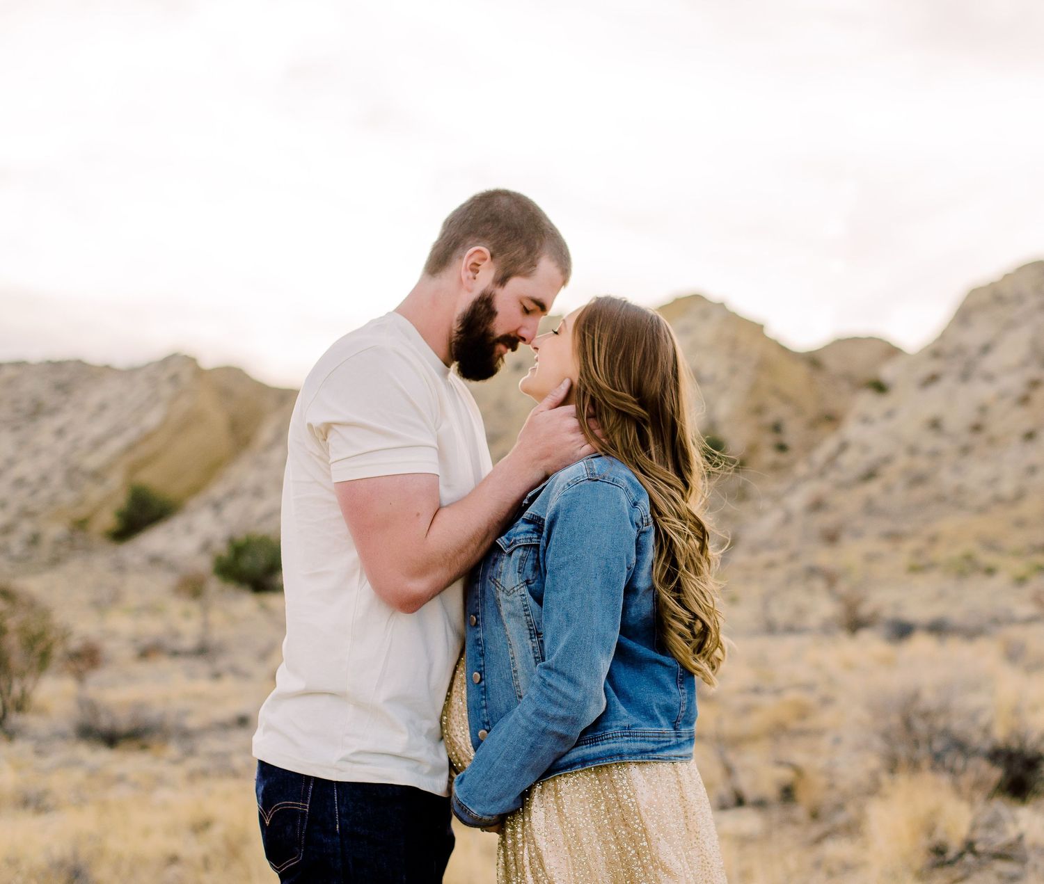 Romantic couple sharing a tender moment in a desert landscape, man in white tee and woman in denim jacket.