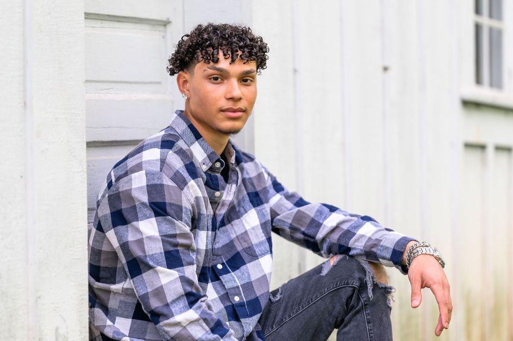 A person wearing a plaid flannel shirt sits casually against a white wall.