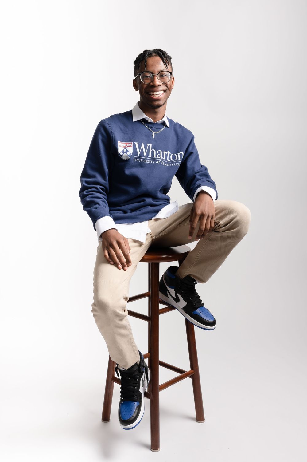 Student wearing Wharton sweatshirt sitting casually on wooden stool against white background.