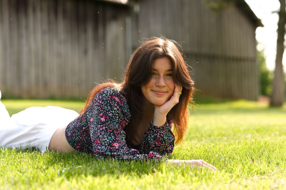 A person with dark hair lies on grass wearing a floral print dress on a sunny day.