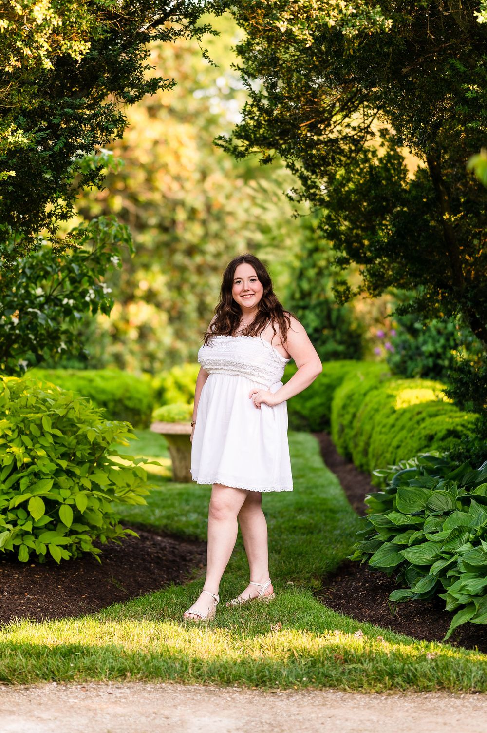 A person in a white summer dress stands on a garden path during golden hour.