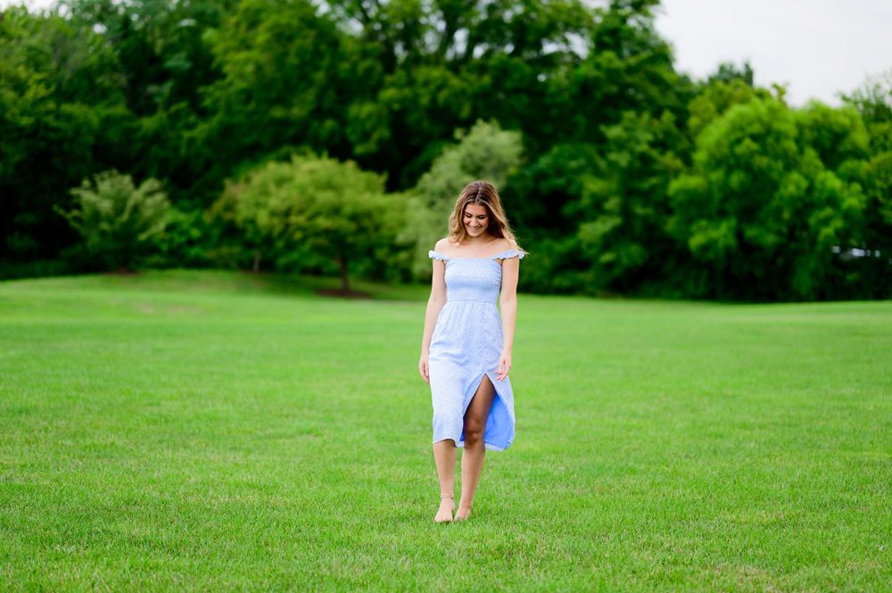 A person in a light blue dress walks barefoot across a lush green lawn with trees in the background.