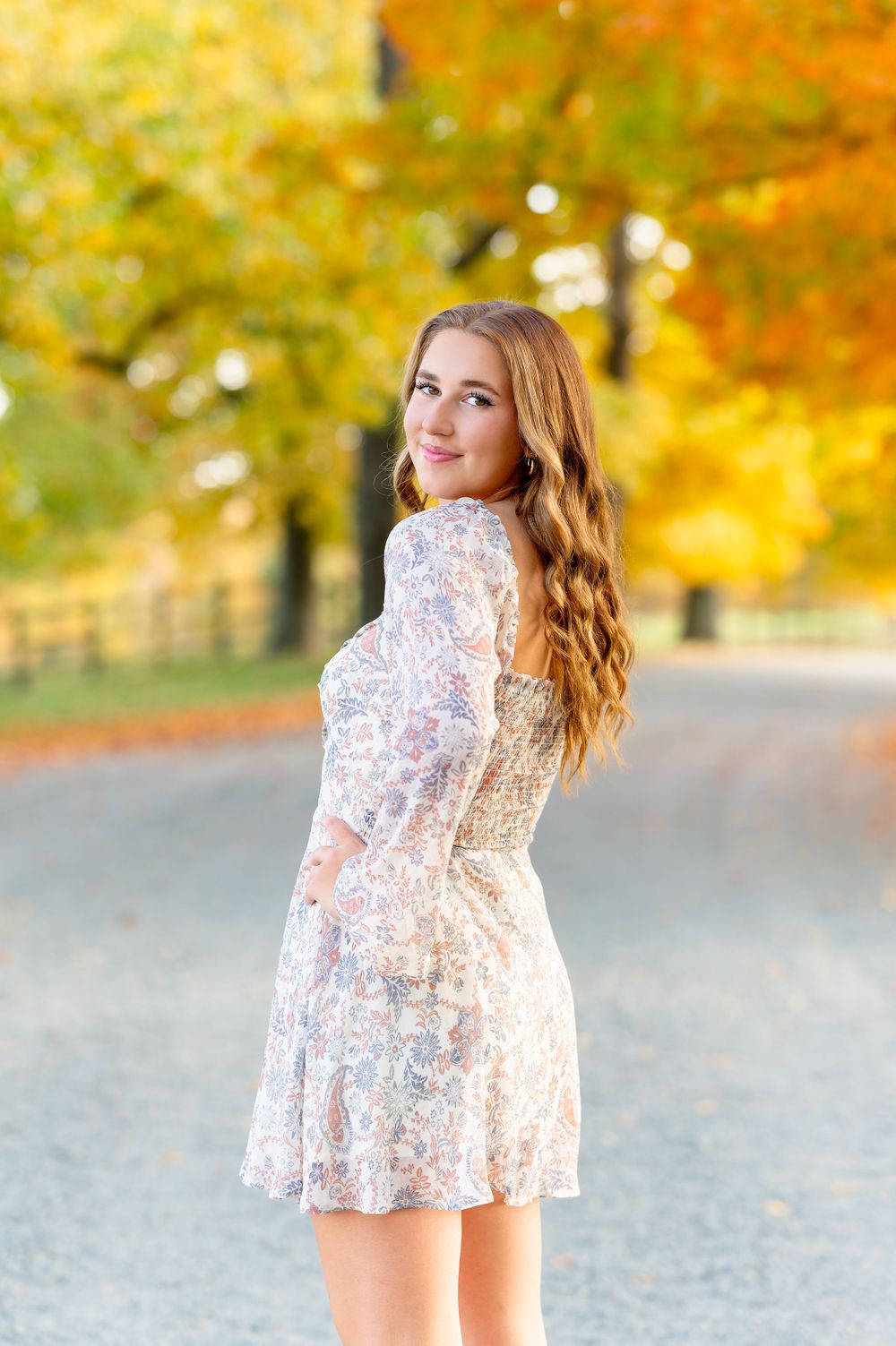 Person in a white lace dress poses on a path with vibrant fall foliage in the background.