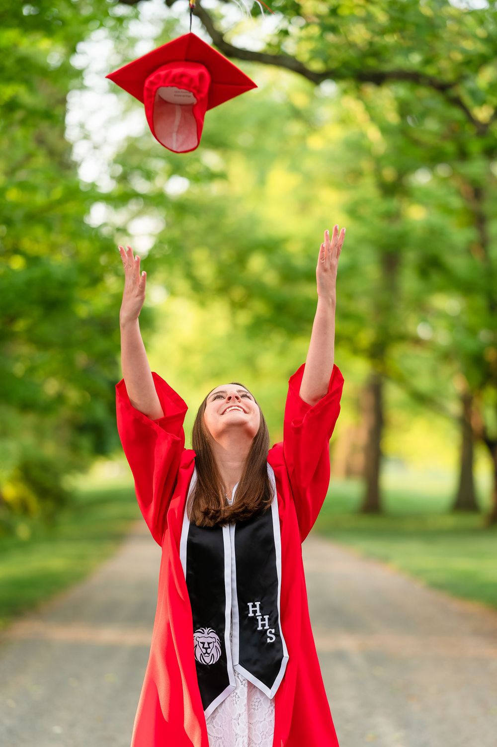 Graduate in red cap and gown tossing graduation cap in the air on tree-lined path.