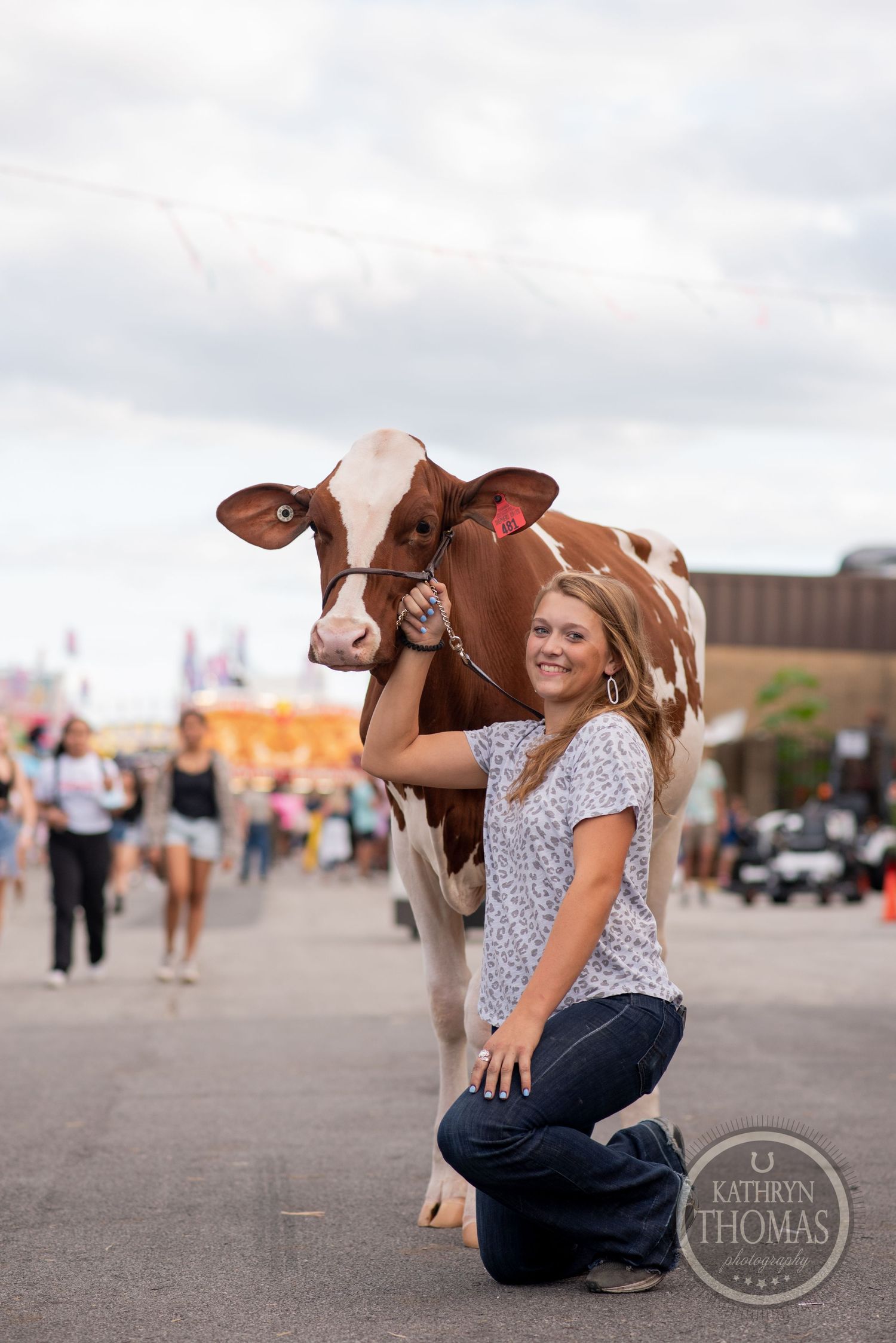 A brown and white dairy cow poses with a person in a floral top at an outdoor fair or event.