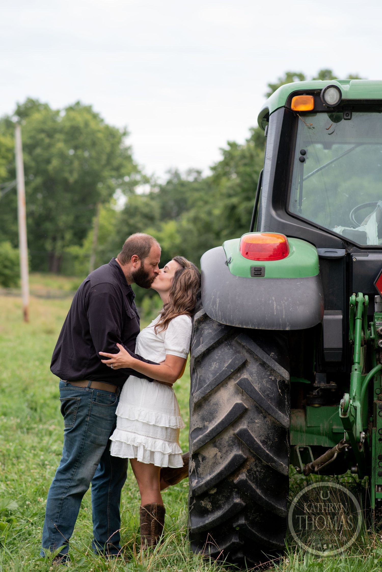 A couple shares an intimate moment beside a large green tractor in a rural field.