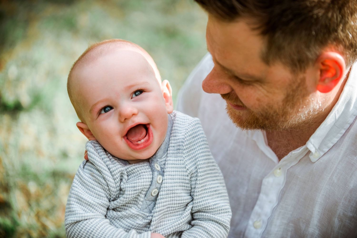 Baby wearing grey sweater laughs with pure joy while being held outdoors.
