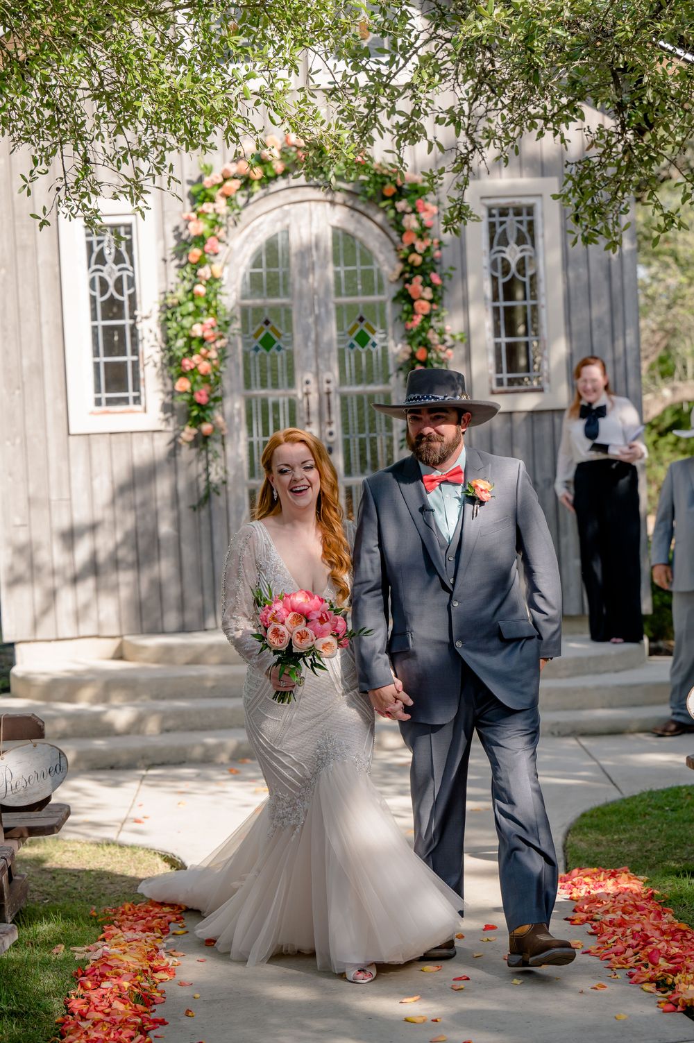 Newlyweds exit chapel under floral arch with scattered rose petals along pathway.