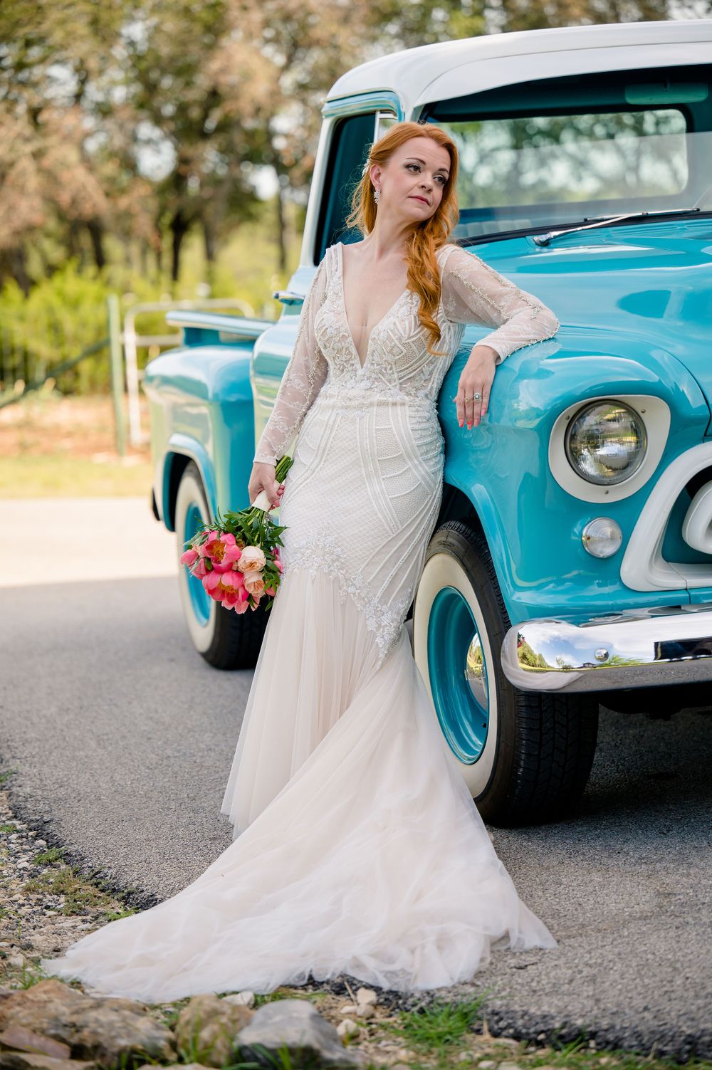 A bride in a lace wedding gown poses beside a classic turquoise pickup truck with a colorful bouquet.