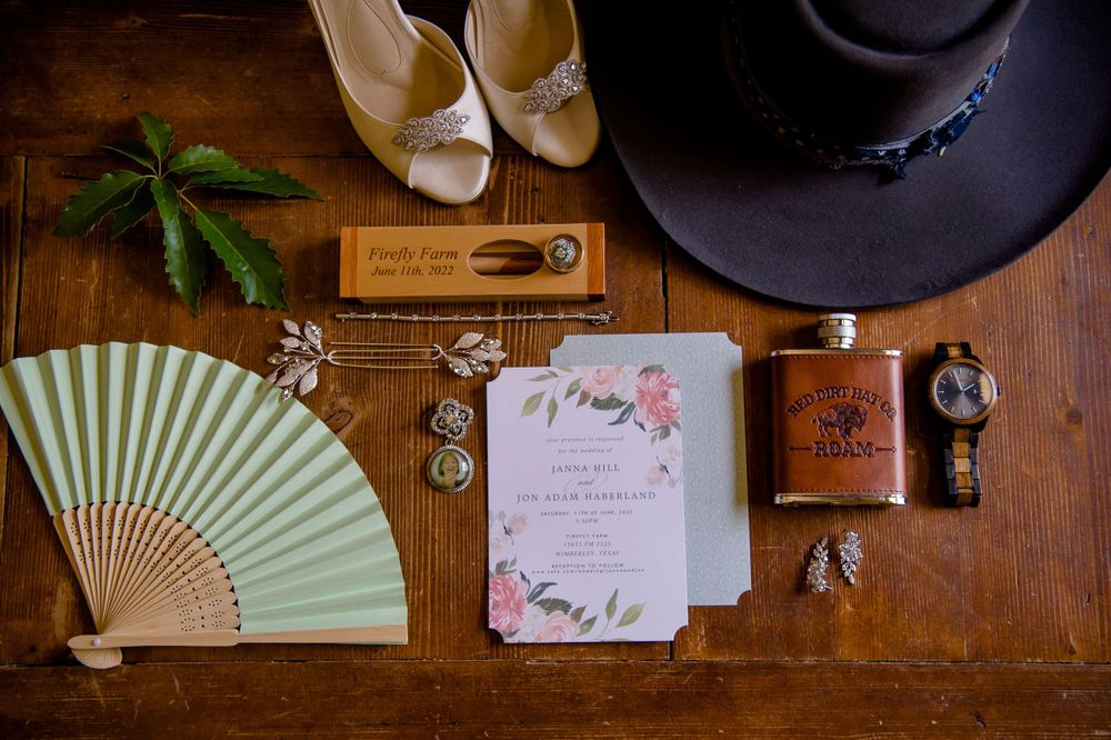 Wedding day details including a mint green fan, floral invitation, high heels, cowboy hat, and hip flask on wooden surface.