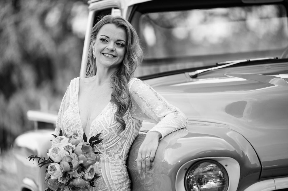 Black and white portrait of a bride leaning against a vintage car with her bouquet.
