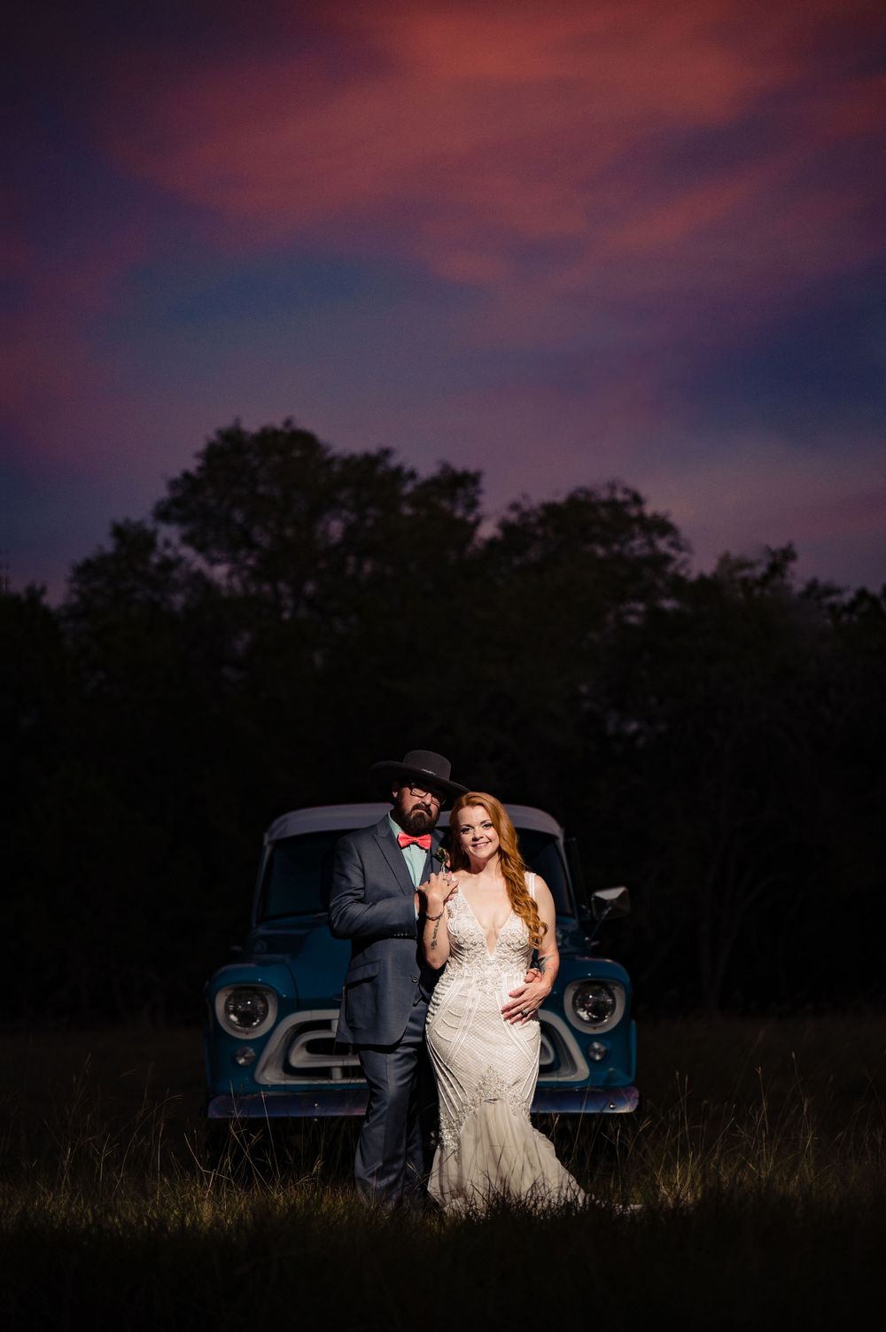 Romantic wedding portrait with vintage blue truck against dramatic sunset sky.