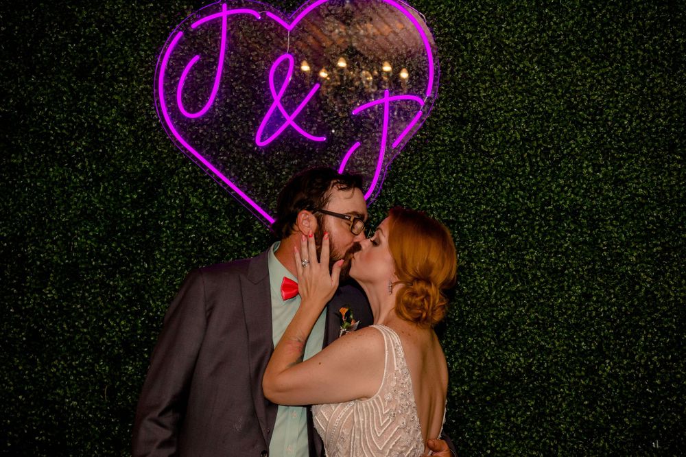 Romantic moment captured under purple neon heart sign against hedge wall backdrop.