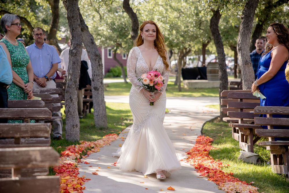 A bride walks down a flower-petal lined aisle in a fitted white lace wedding dress.